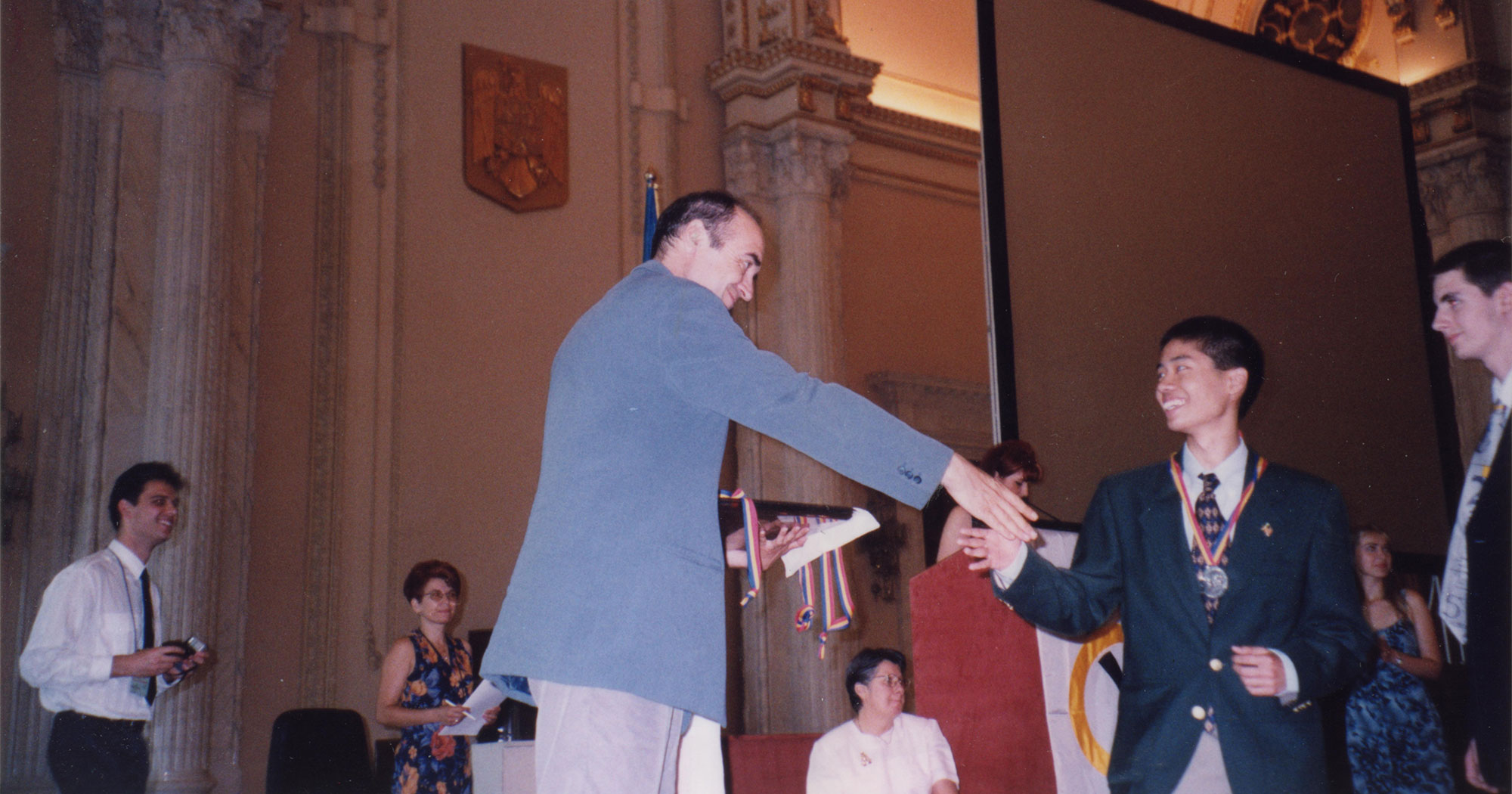 Loh onstage and shaking hands with a medal on his chest at the 1999 IMO in Bucharest, Romania.