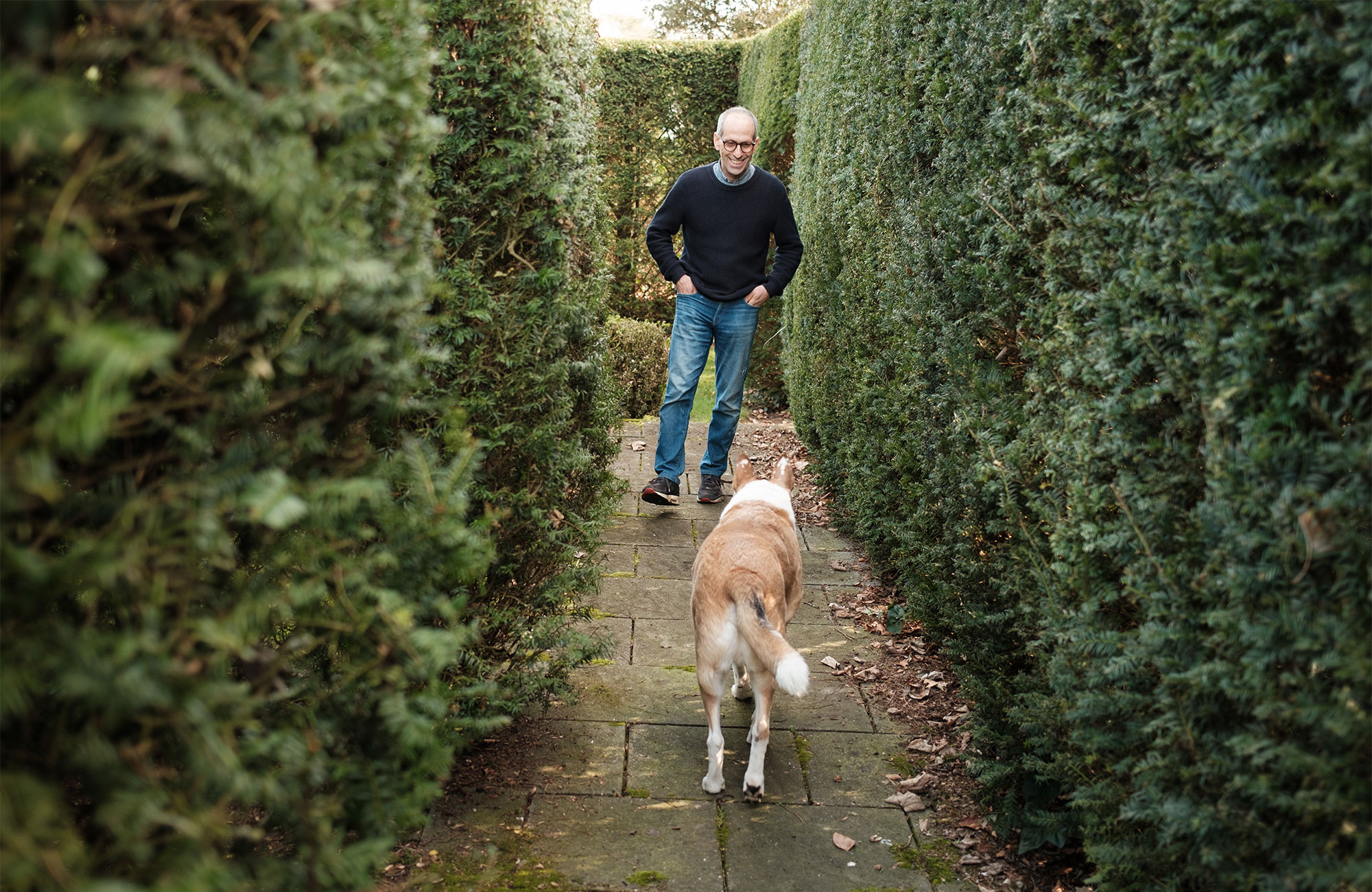 Arik Kershenbaum and his dog standing in a narrow path between tall hedges.