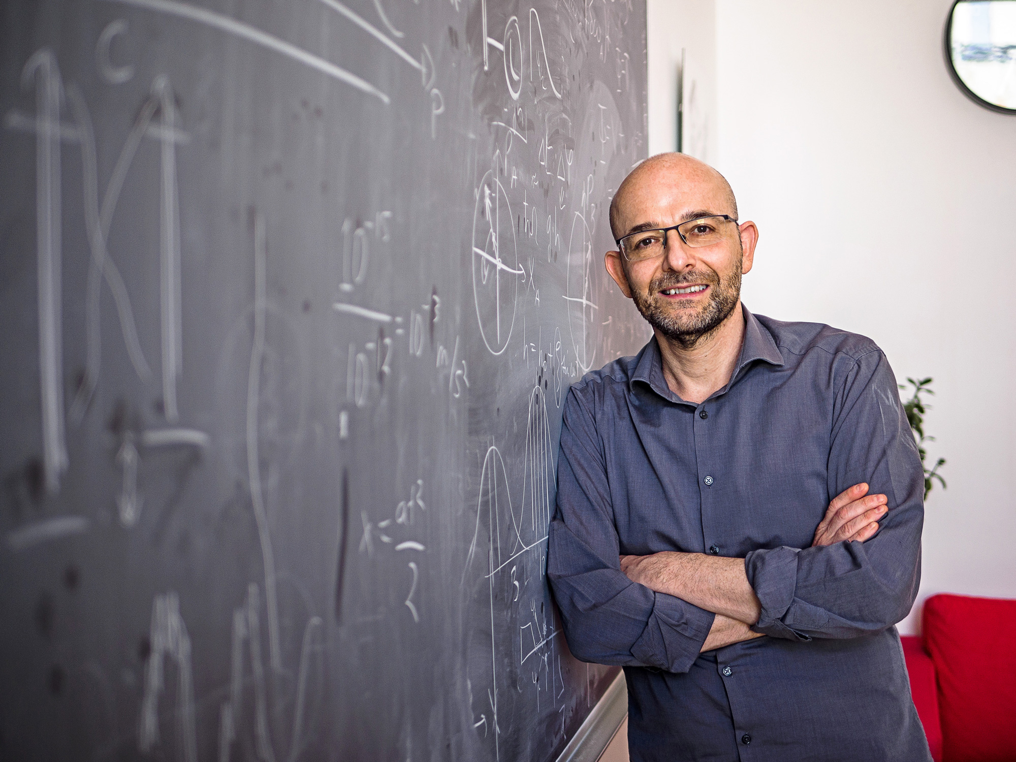 A diptych of two photos; one shows a bald man in glasses with arms crossed leaning against a blackboard and the other shows a woman in a blazer using a marker to point at an equation on a whiteboard.