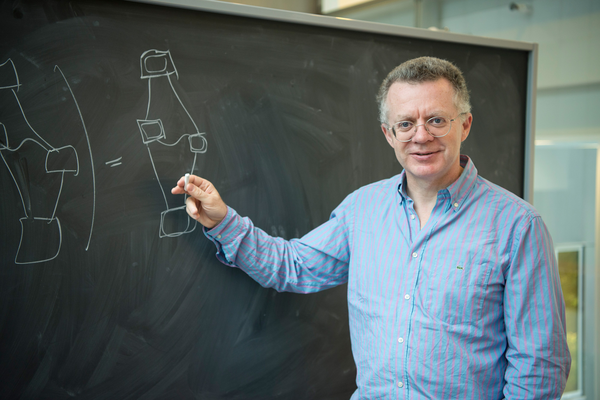 A man with curly gray hair and wireframe glasses smiles for the camera while holding a piece of chalk up to a blackboard.