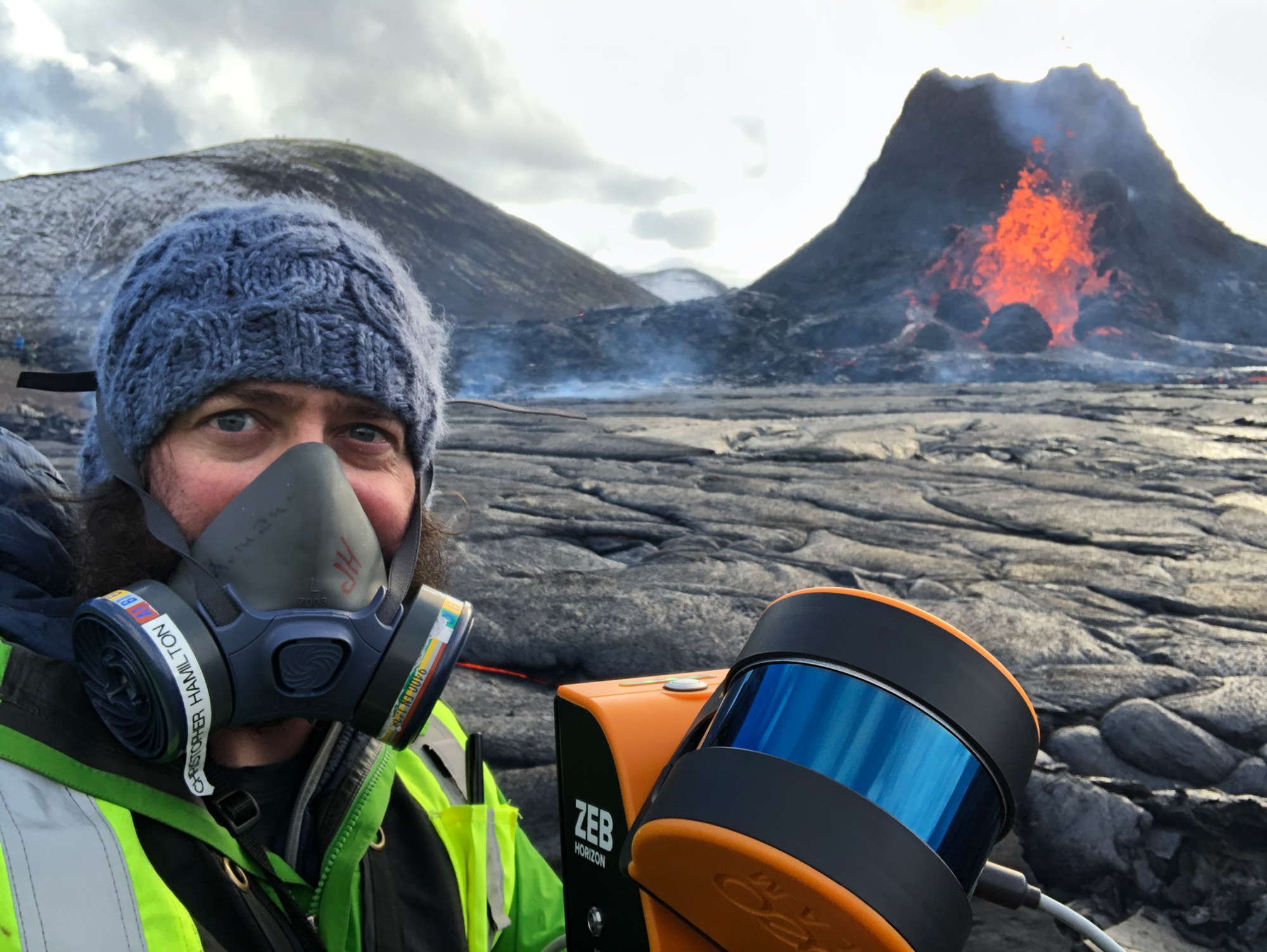 A man in a mask holding a LIDAR instrument in front of a volcano.