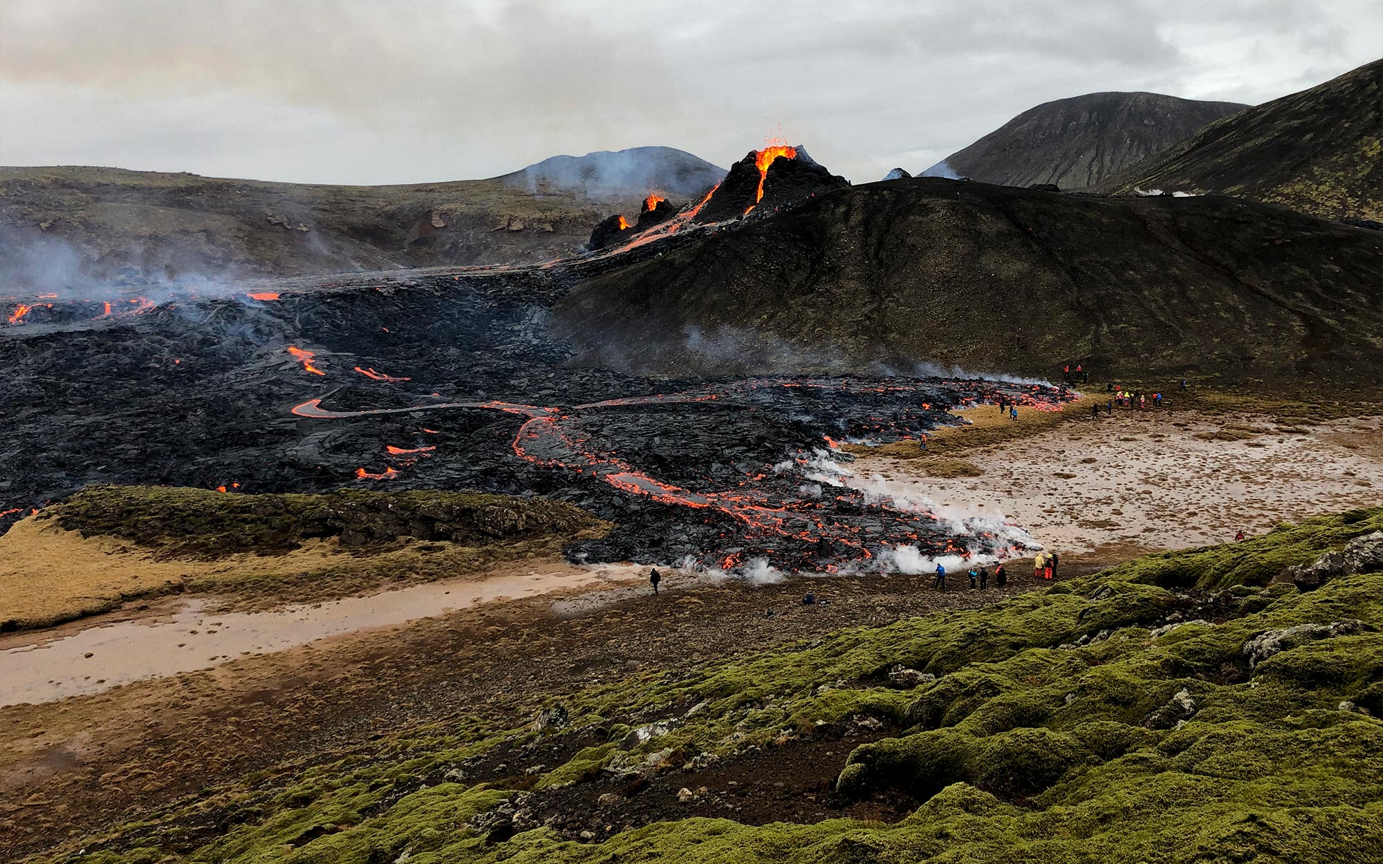 A volcano with orange lava flows coming down into a valley, with about 20 people standing at the base.