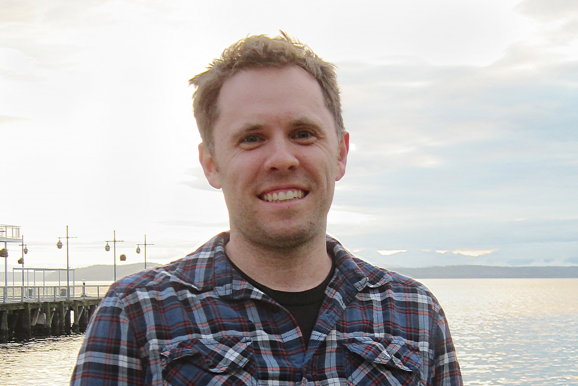 A man with a checked shirt standing in front of the water. A man with round glasses and a grey shirt.