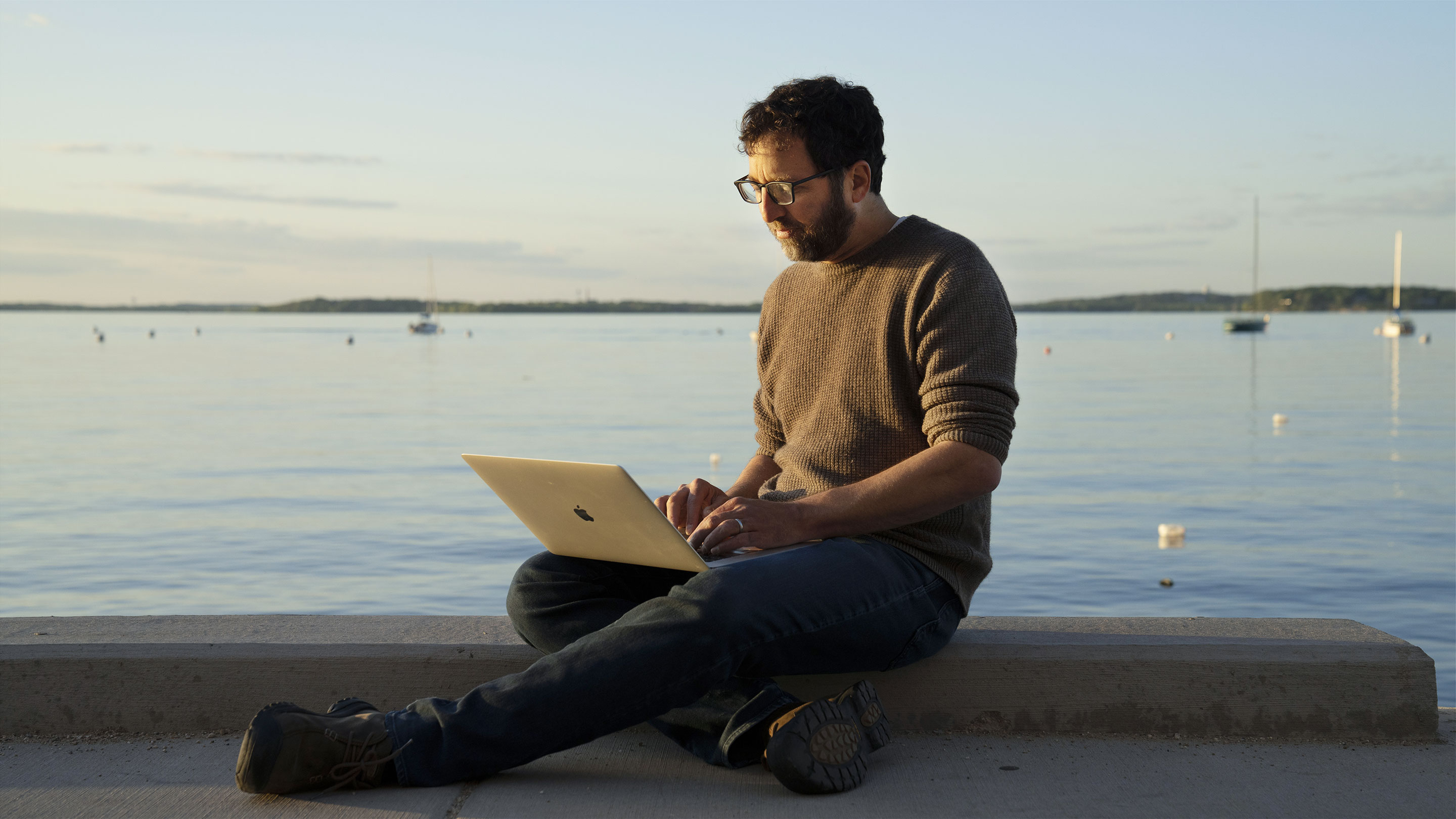 Color photo of Jordan Ellenberg sitting with a laptop by a lake at sunset