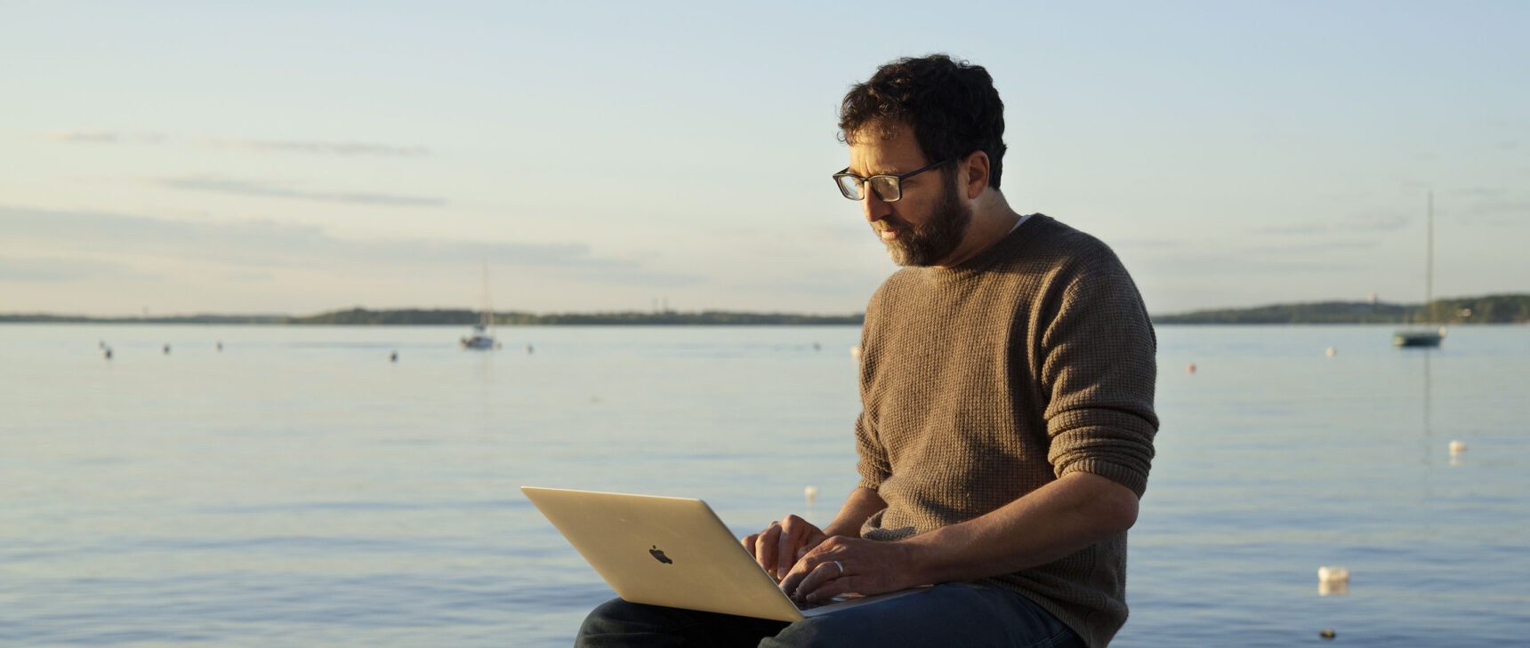 Color photo of Jordan Ellenberg sitting with a laptop by a lake at sunset