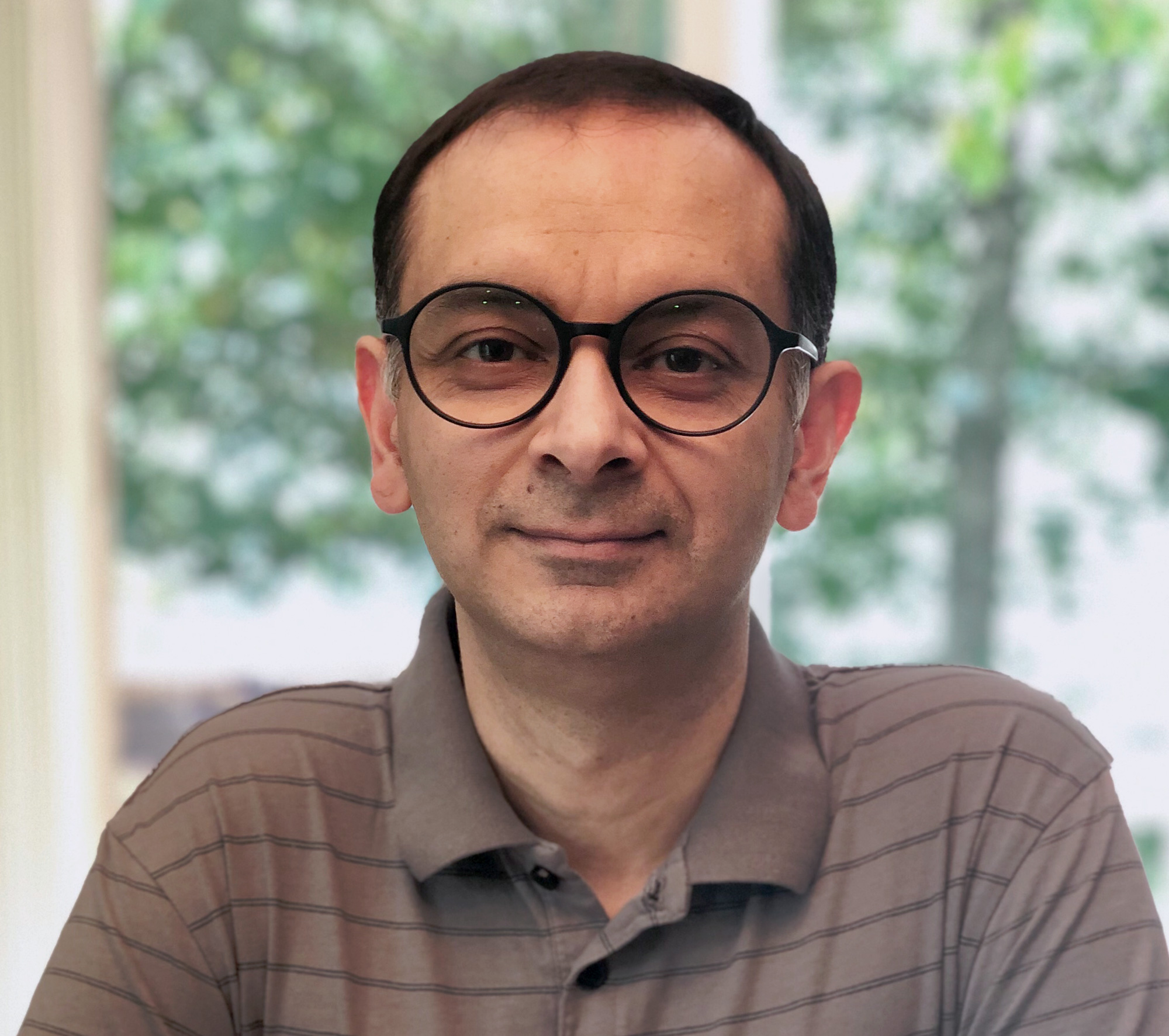 A man with a checked shirt standing in front of the water. A man with round glasses and a grey shirt.