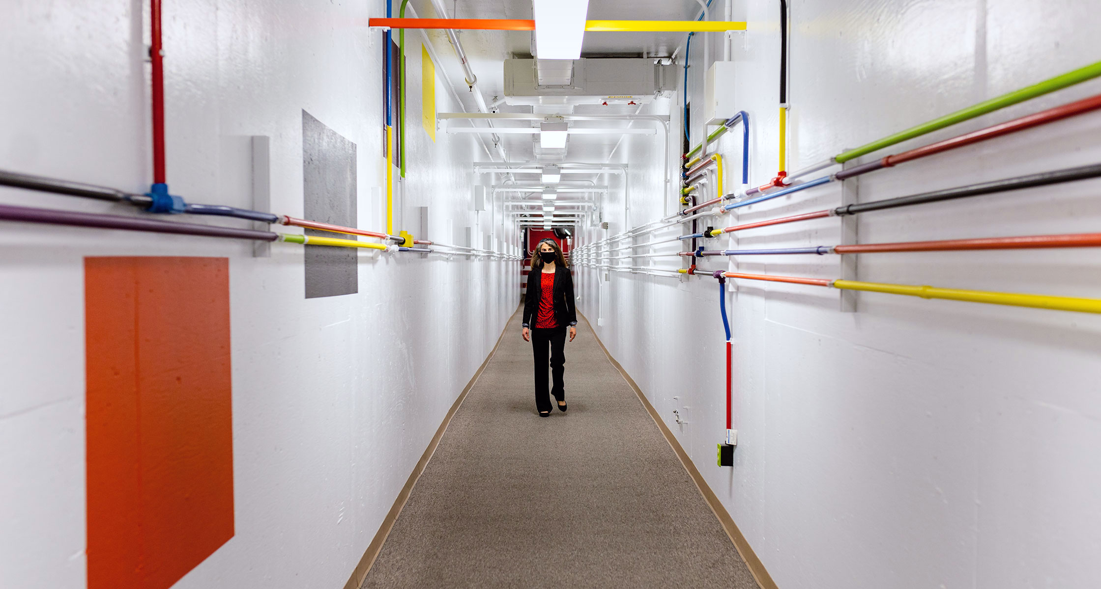 A woman walks down a white corridor with colorful pipes running down the walls.