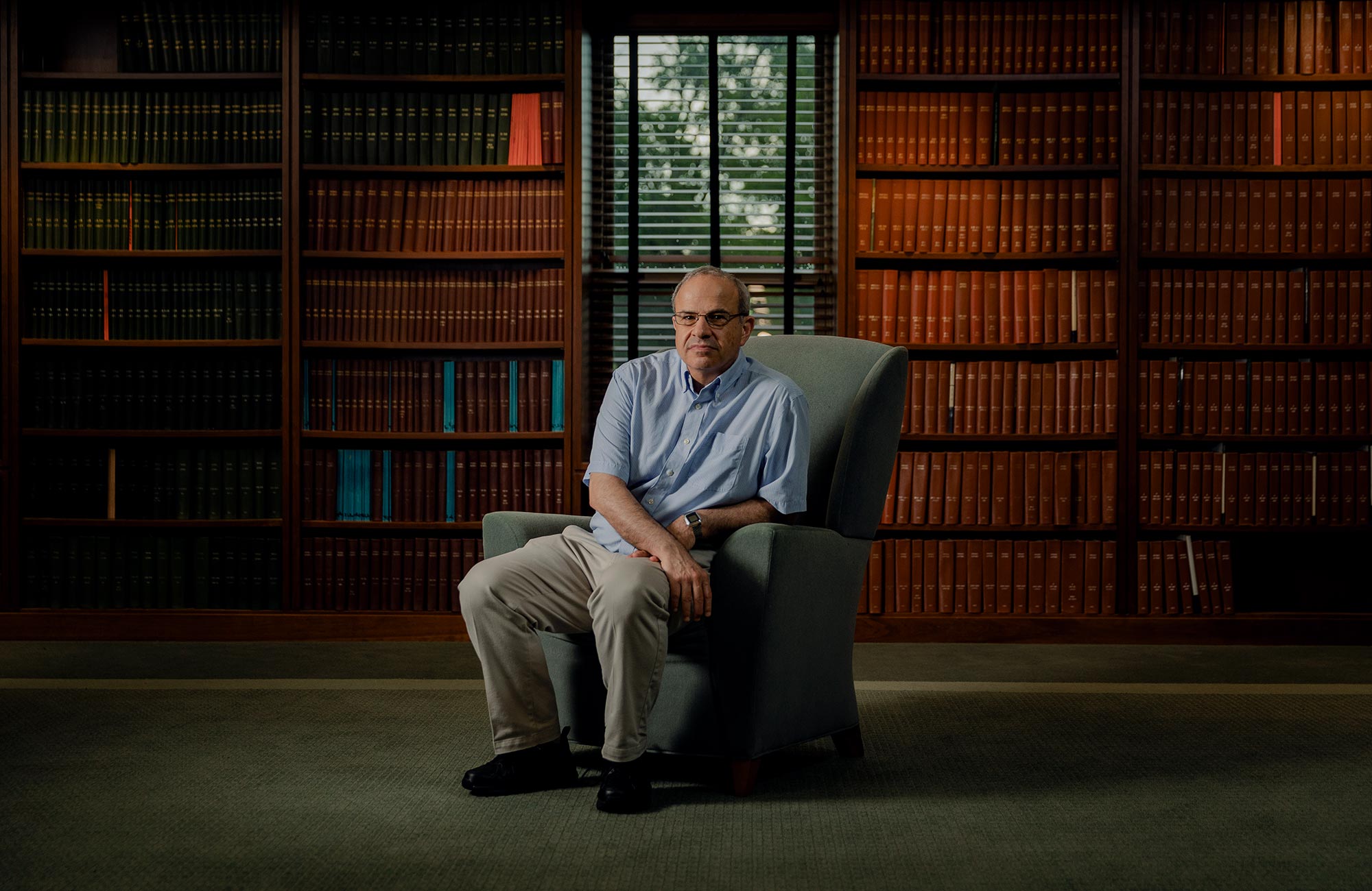 Color photo of Seiberg sitting in an easy chair in front of multiple shelves full of books.