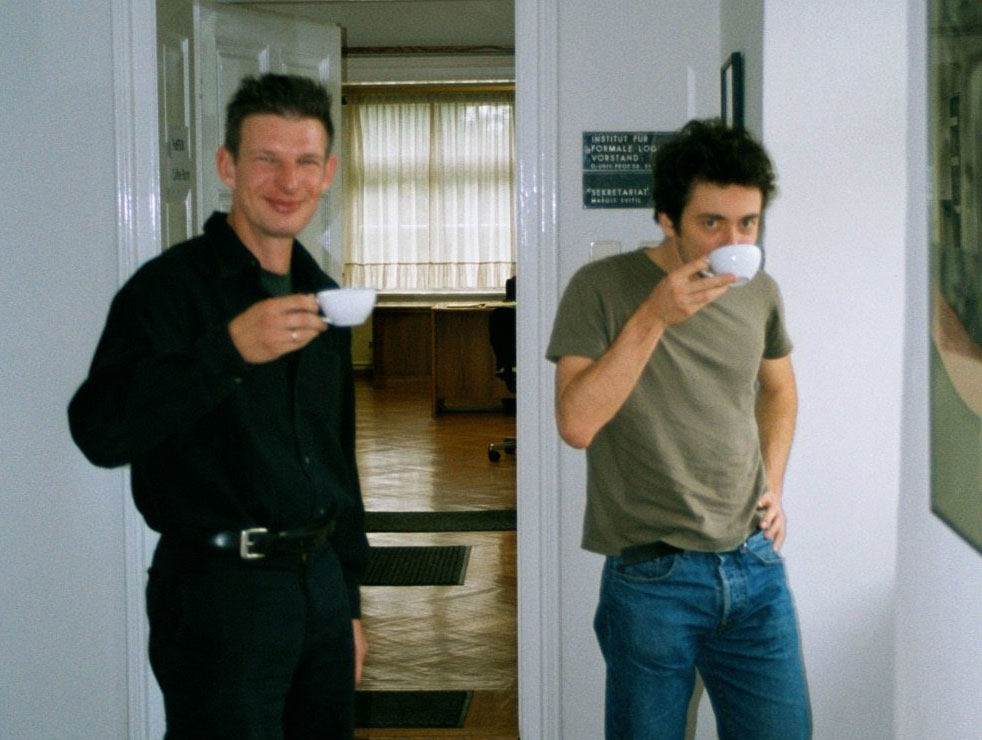 Two men standing up and drinking from coffee cups.