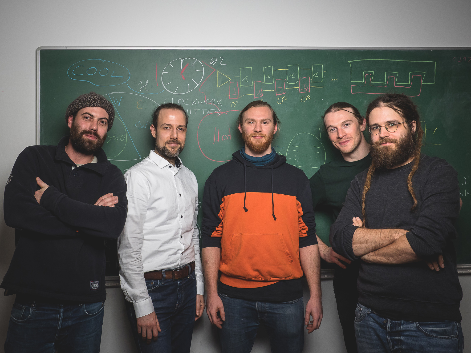 Five men posing in front of a blackboard that displays sketches related to clocks.