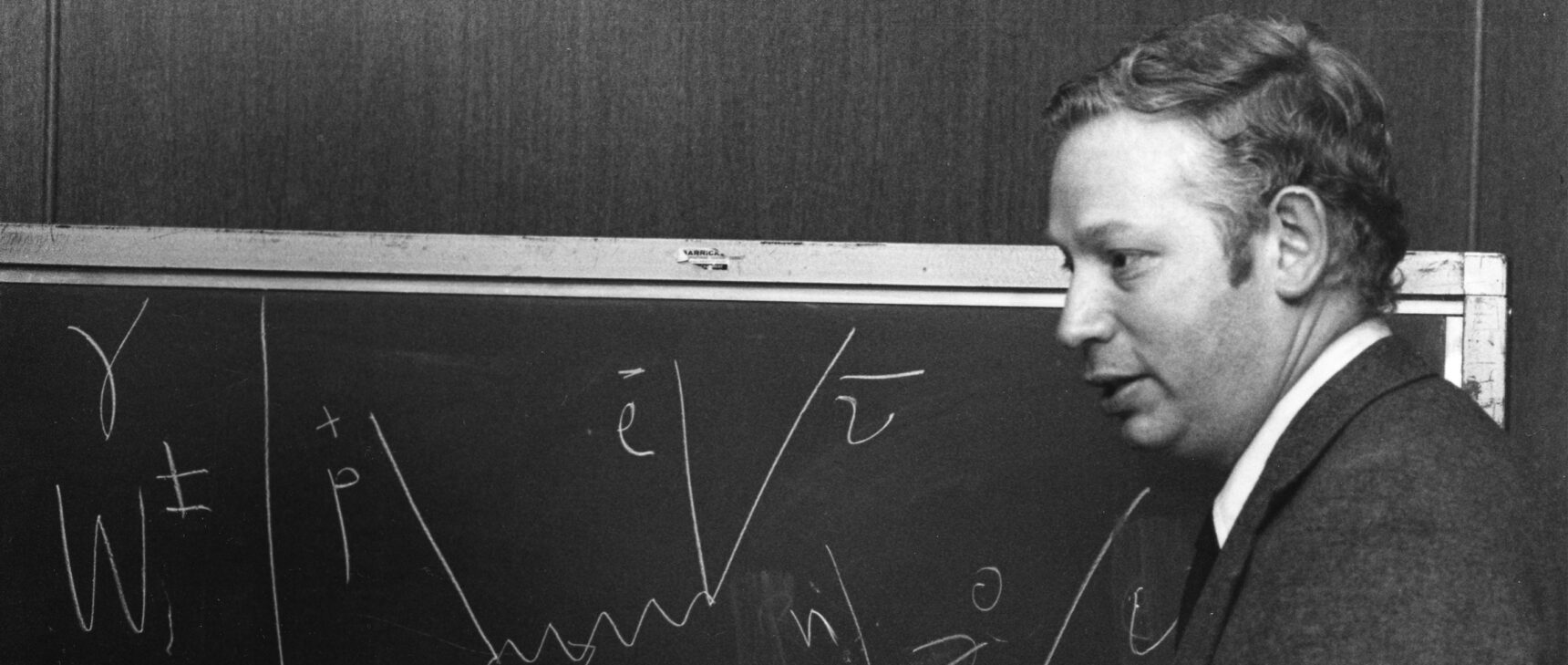 Black and white photo of Steven Weinberg in front of a chalkboard showing particle interactions