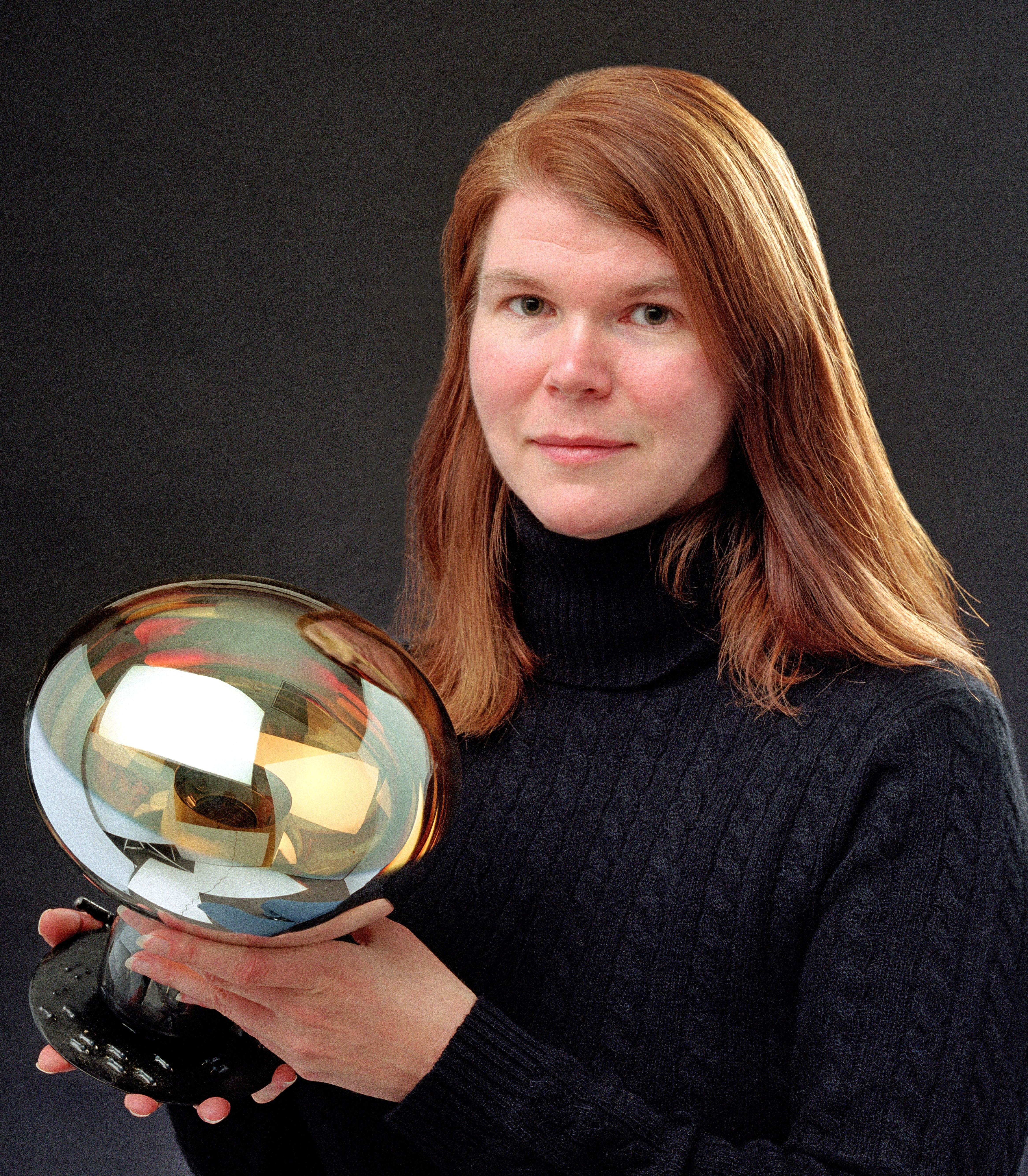 Janet Conrad, a woman with red hair in a black turtleneck, poses with a round, shiny object.