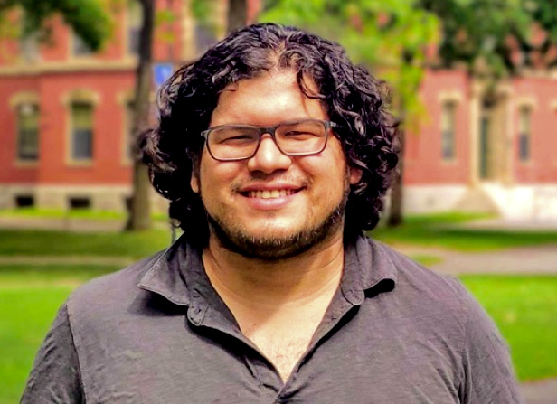 Photo portrait of Carlos Argüelles-Delgado, a smiling man with glasses and curly black hair, standing on a college campus.