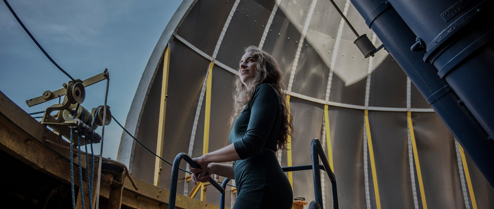 Kreidberg standing inside the dome of an observatory. Two tubes of a dual refractor telescope point through an opening in the dome.