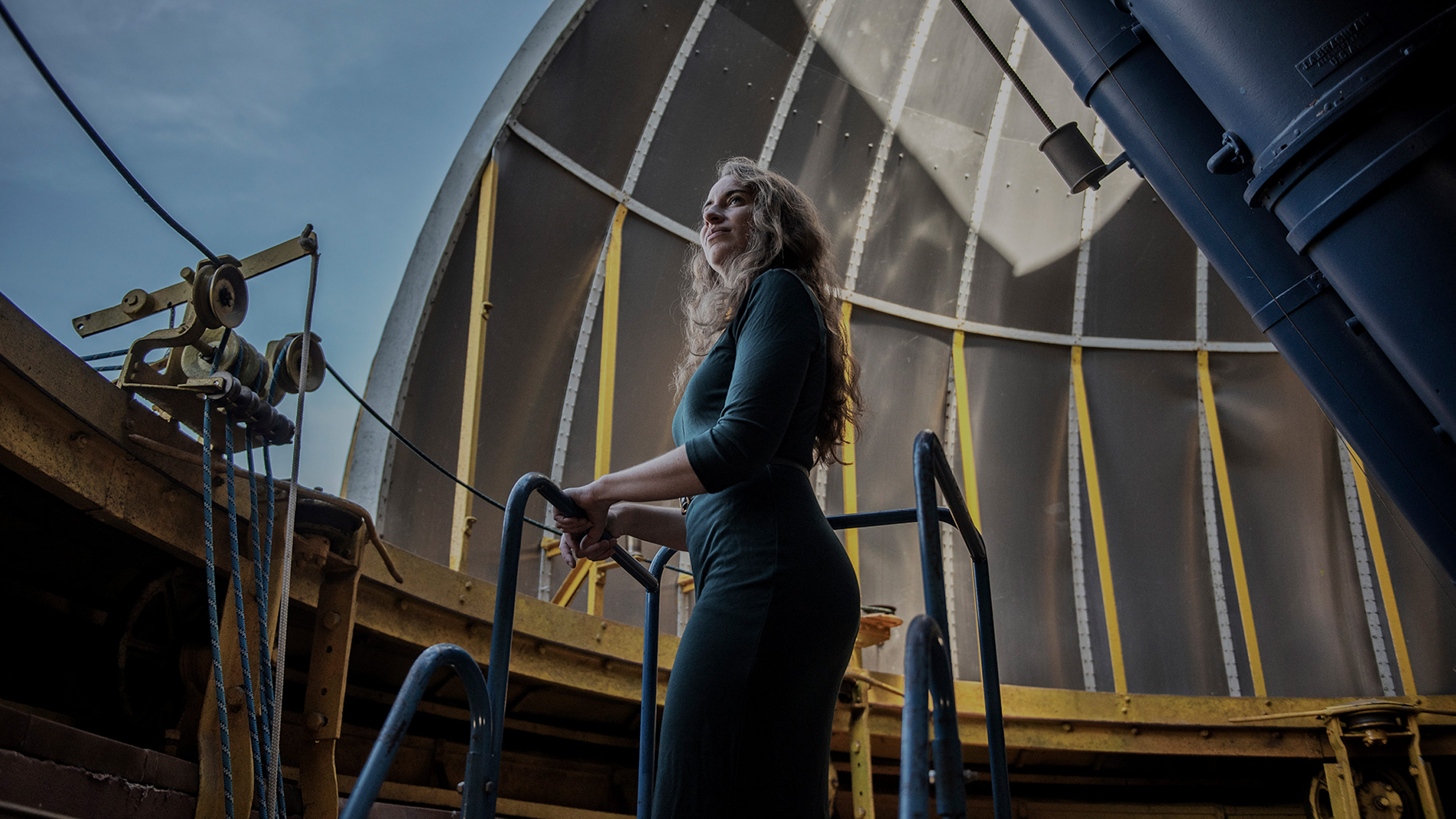 Kreidberg standing inside the dome of an observatory. Two tubes of a dual refractor telescope point through an opening in the dome.