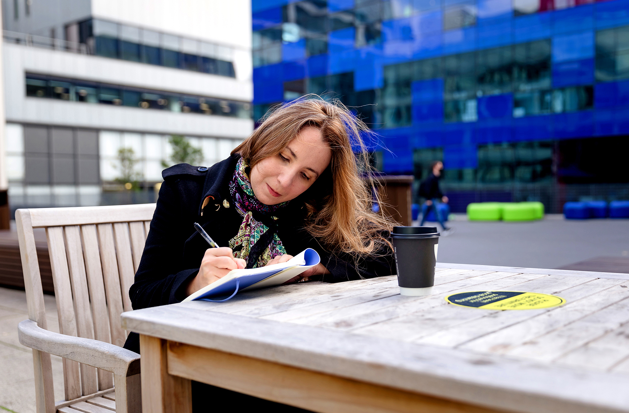 Outdoor photo of Ana Caraini sitting at a wooden table with a blue building in the background