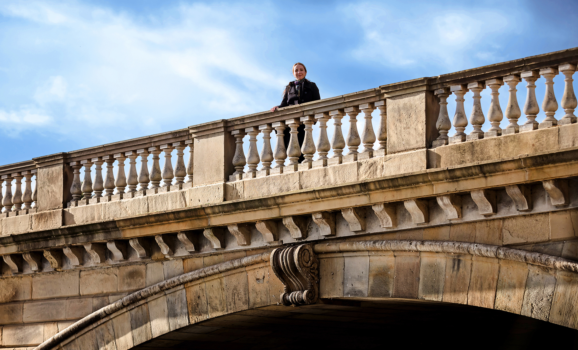 Color photo of Ana Caraiani on a bridge with a partly cloudy sky in the background