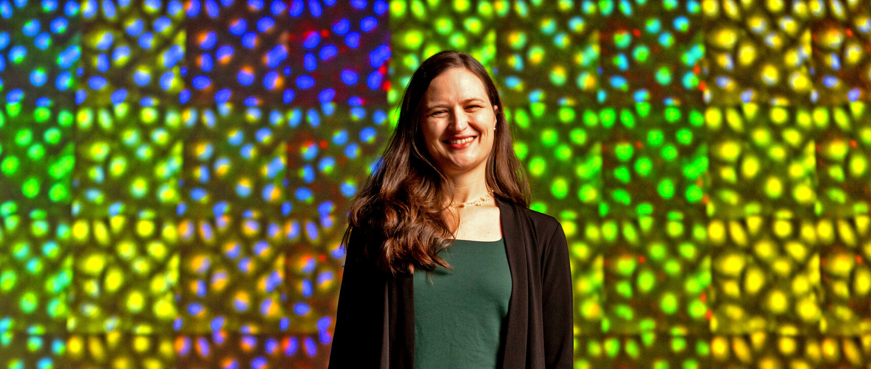 Photo of Anne Carpenter of the Broad Institute standing in front of a wall of colored microscopy images.