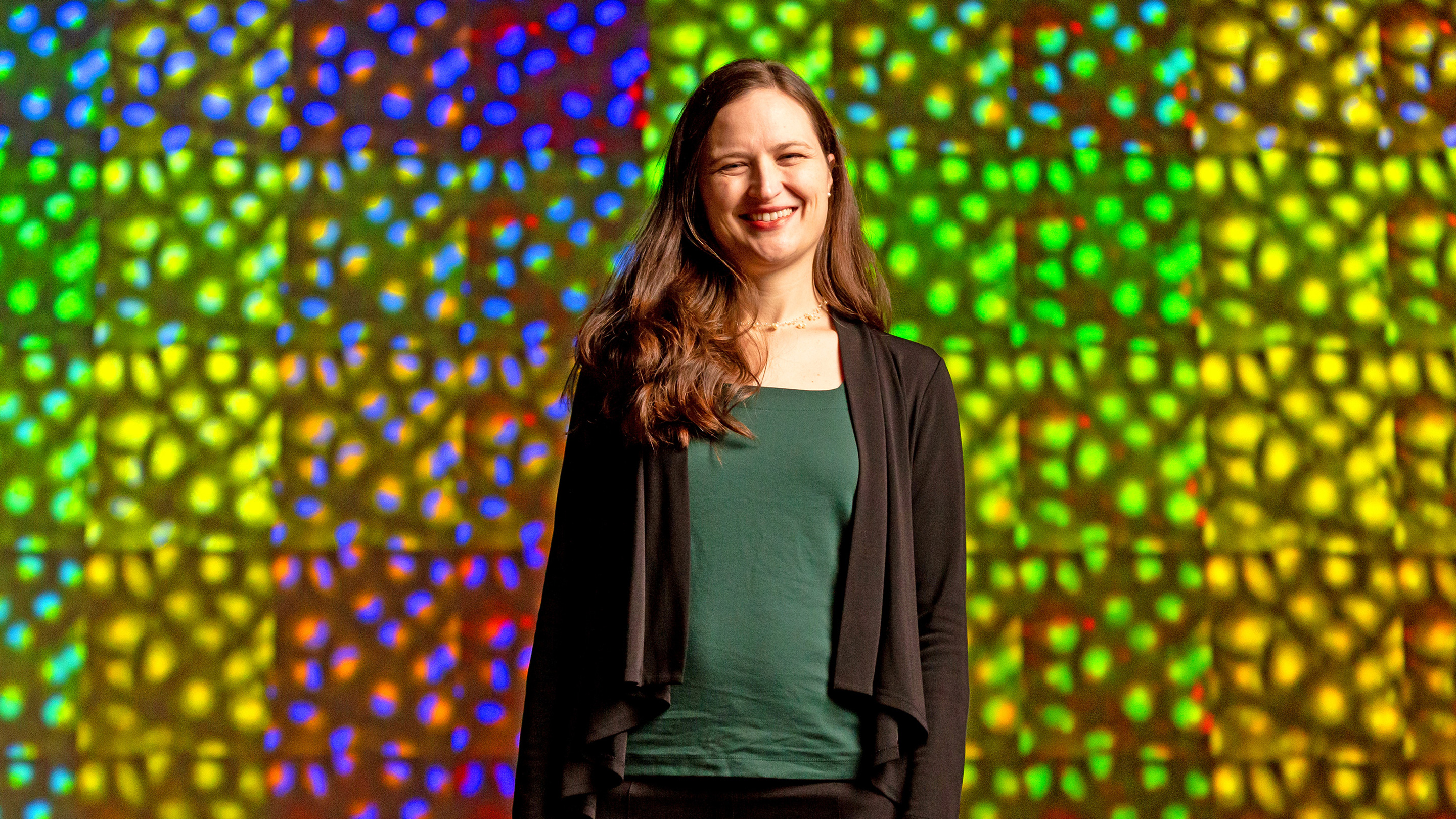 Photo of Anne Carpenter of the Broad Institute standing in front of a wall of colored microscopy images.