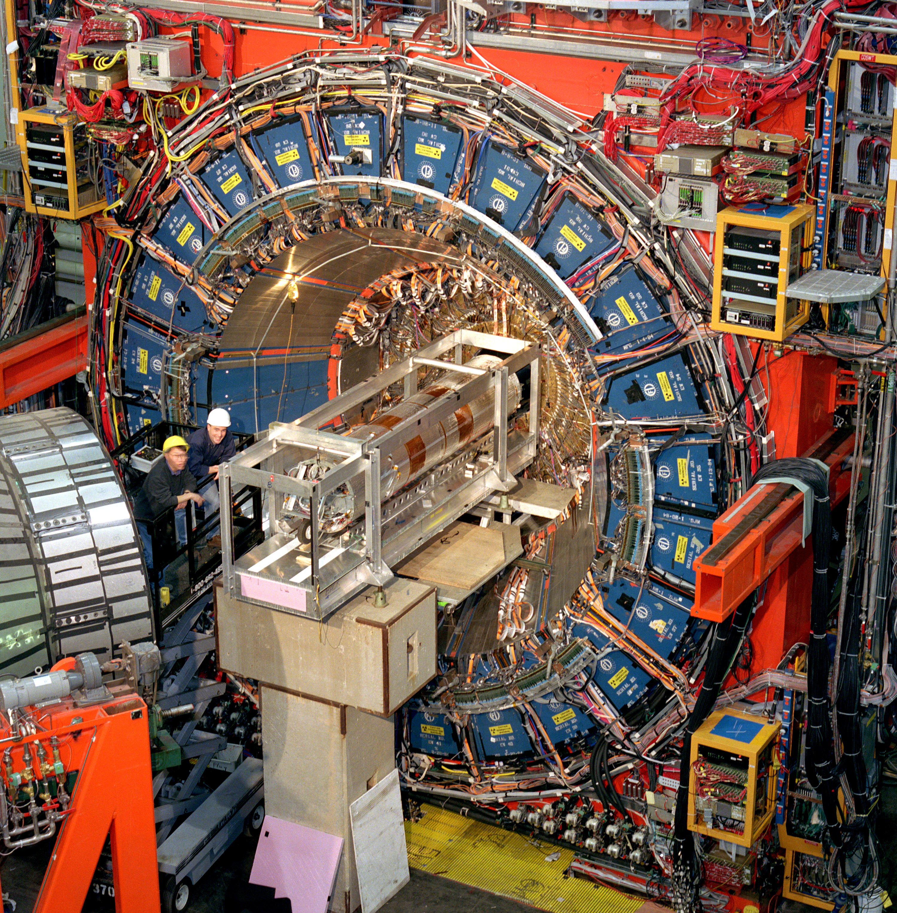 Photo of a large cylindrical contraption. Two people in hard hats stand on a scissor lift in front of the detector.]