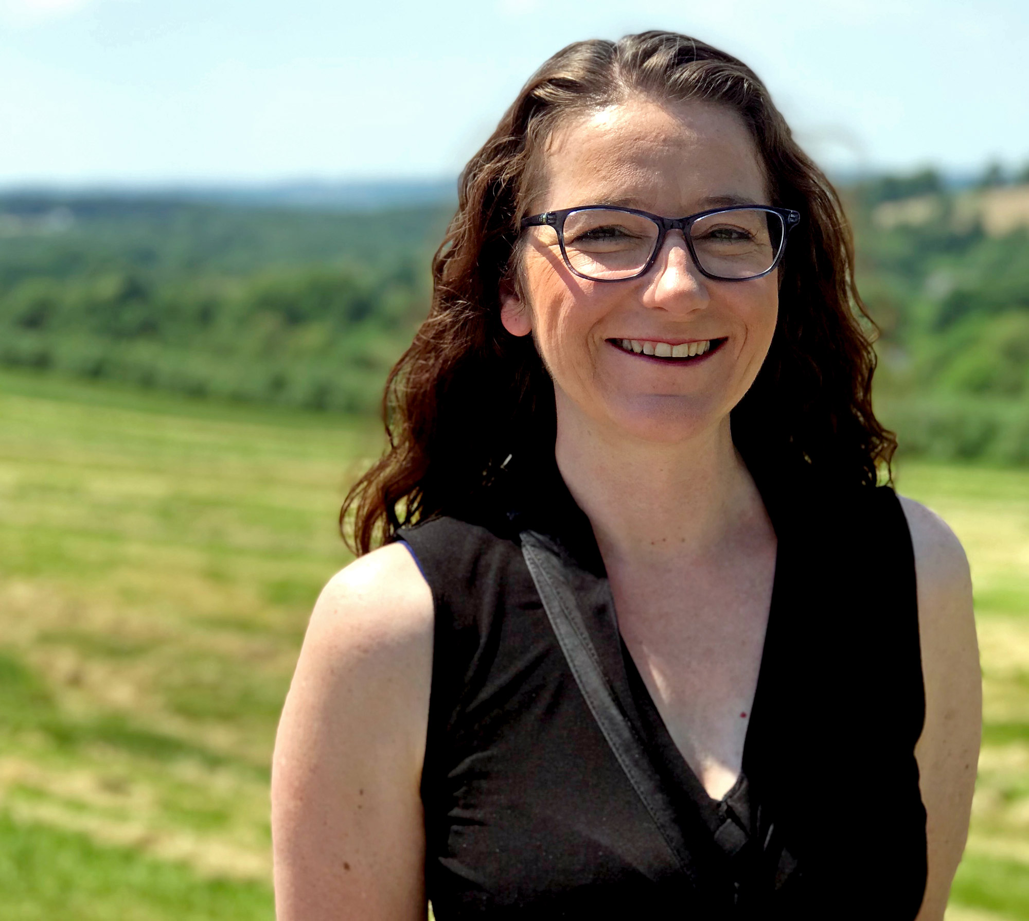 A woman with dark hair and glasses stands in a field smiling at the camera.