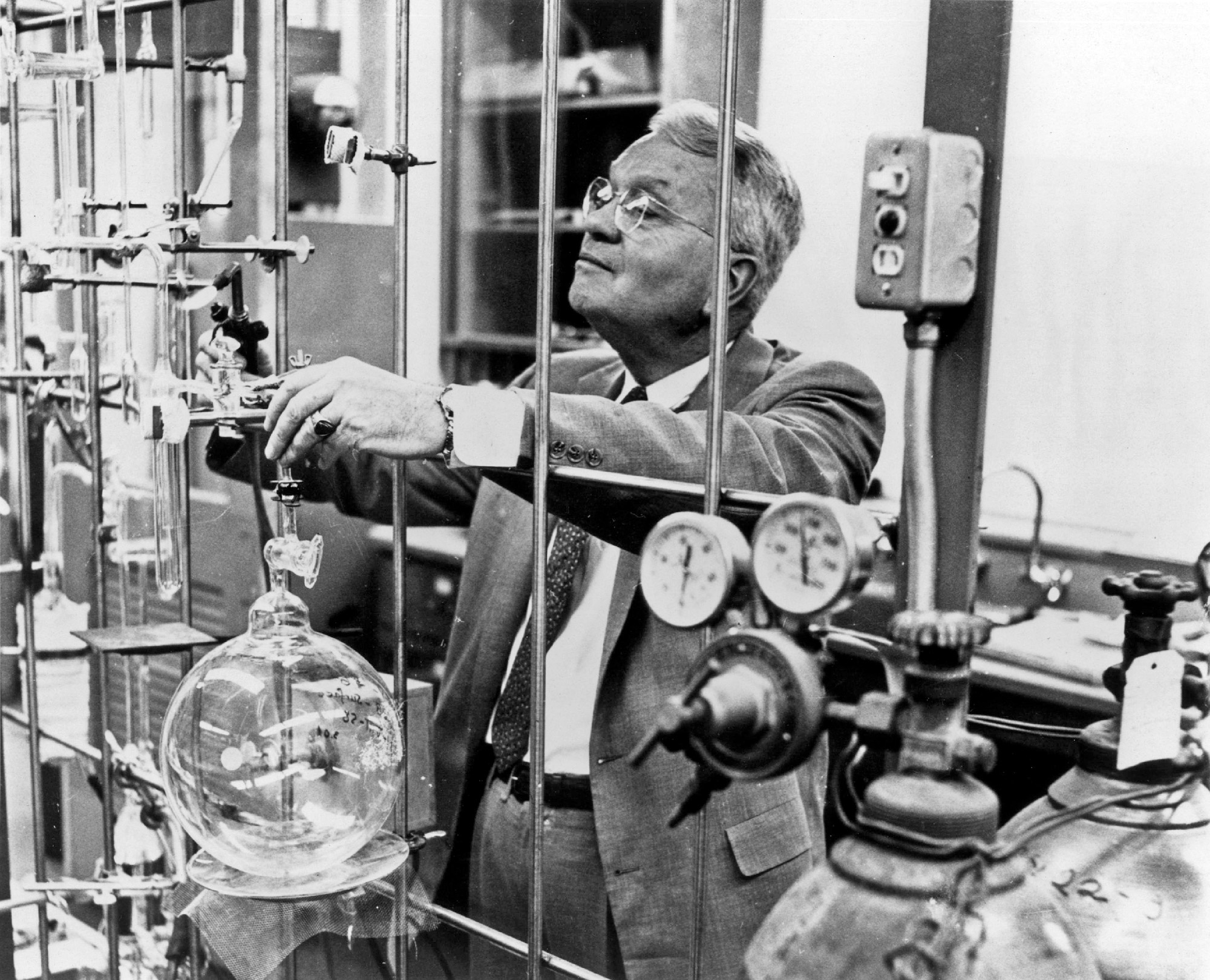 Black-and-white photo of a man in glasses, a suit and tie fiddling with an experiment in a chemistry lab