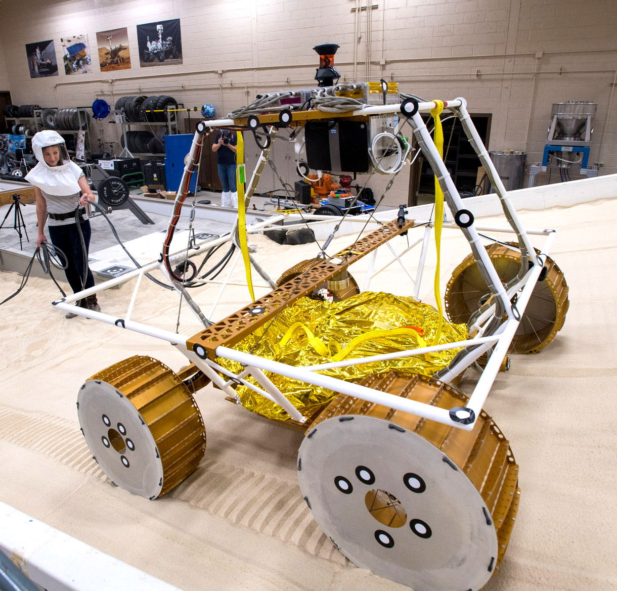 A buggy climbs a sandy incline in a lab while two engineers in shields look on.