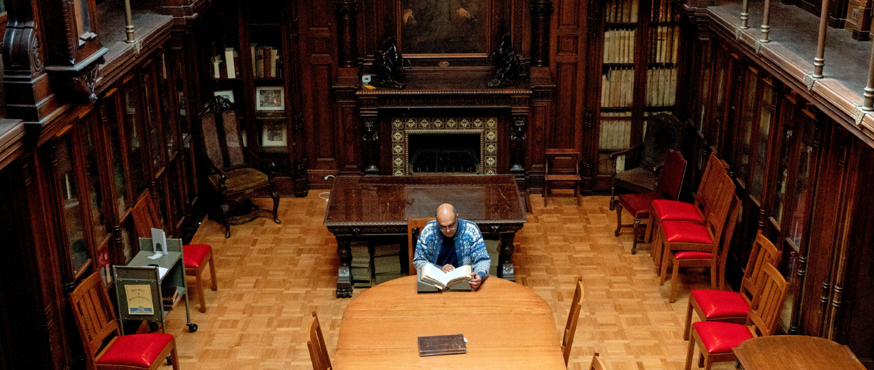 A man sits at a table in the middle of a wood-paneled library, perusing a large book.