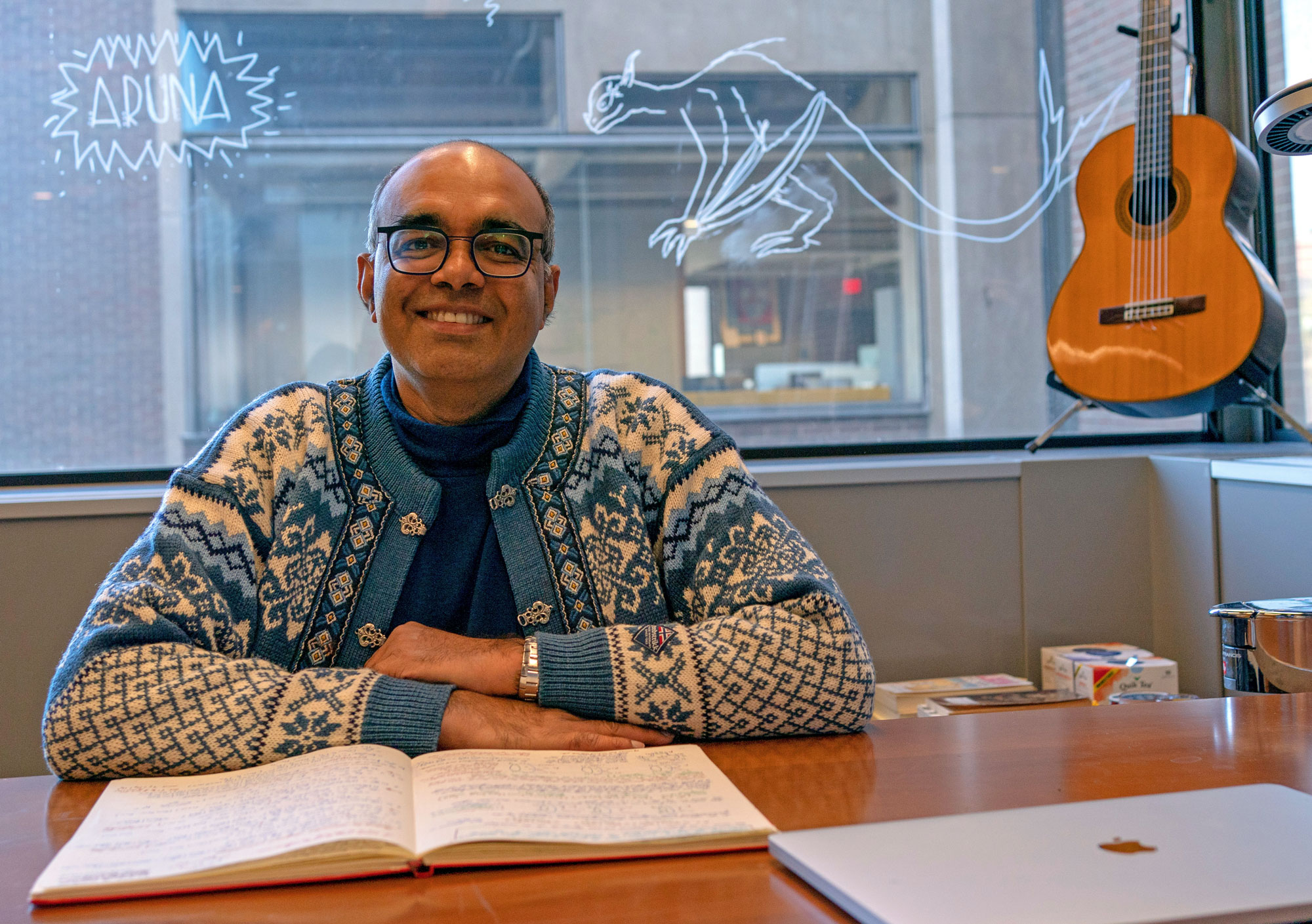 A smiling man in a blue and white wool sweater sits at a desk. A guitar hangs on an adjacent wall.