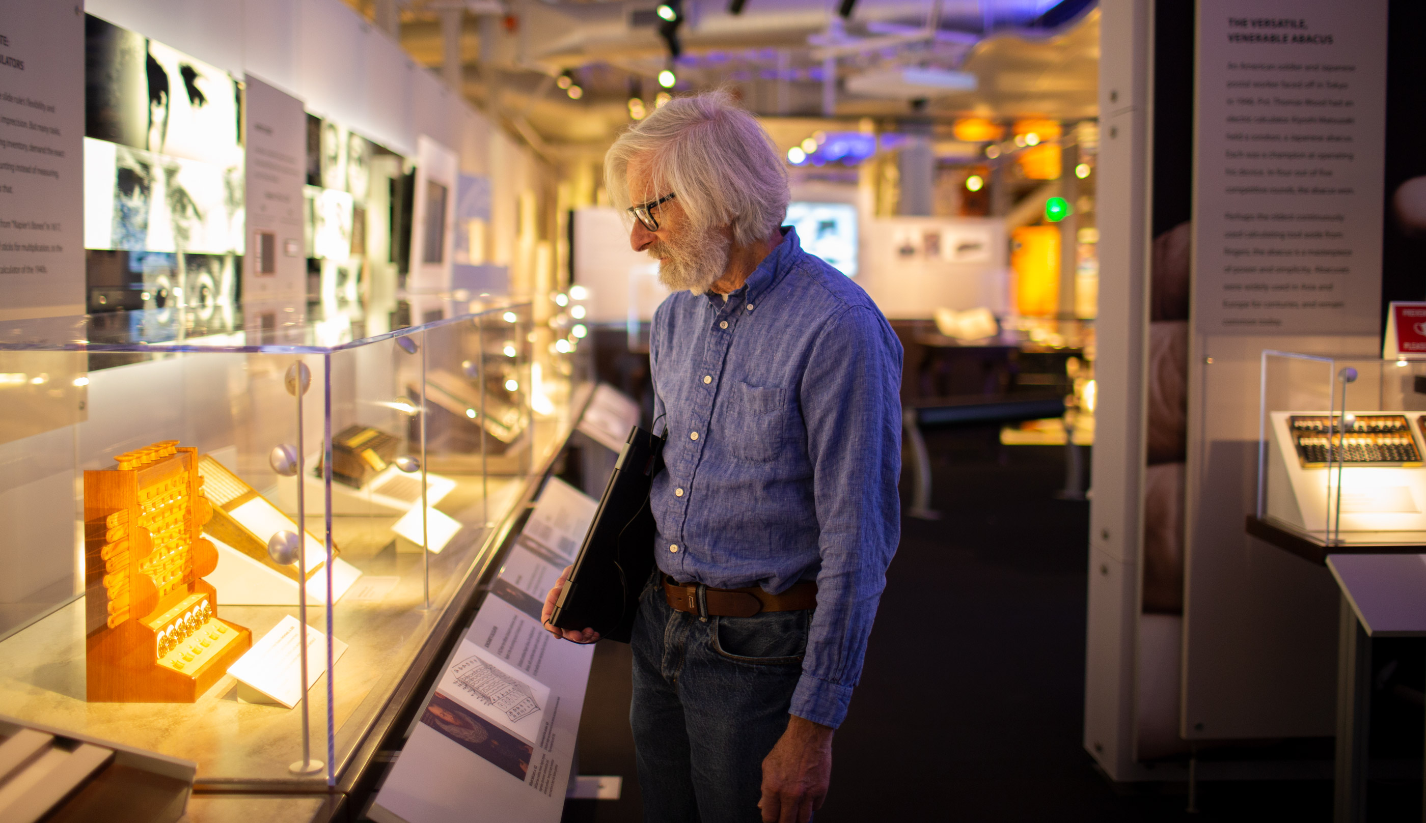 Photo of Leslie Lamport in a blue shirt and jeans standing in front of a brightly lit museum exhibit showing early computers