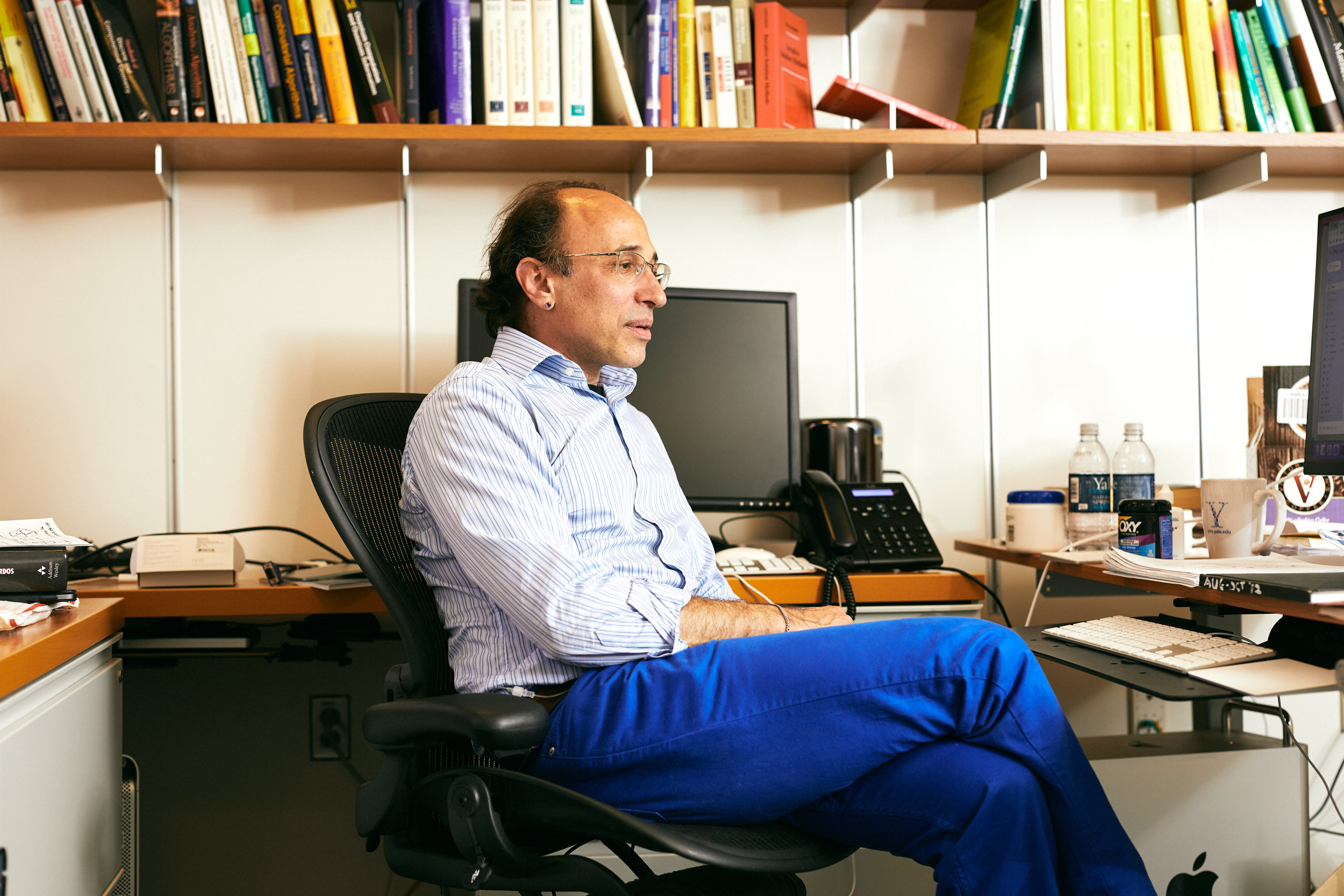 Daniel Spielman sits at a desk with computers, with a colorful row of books on a shelf above