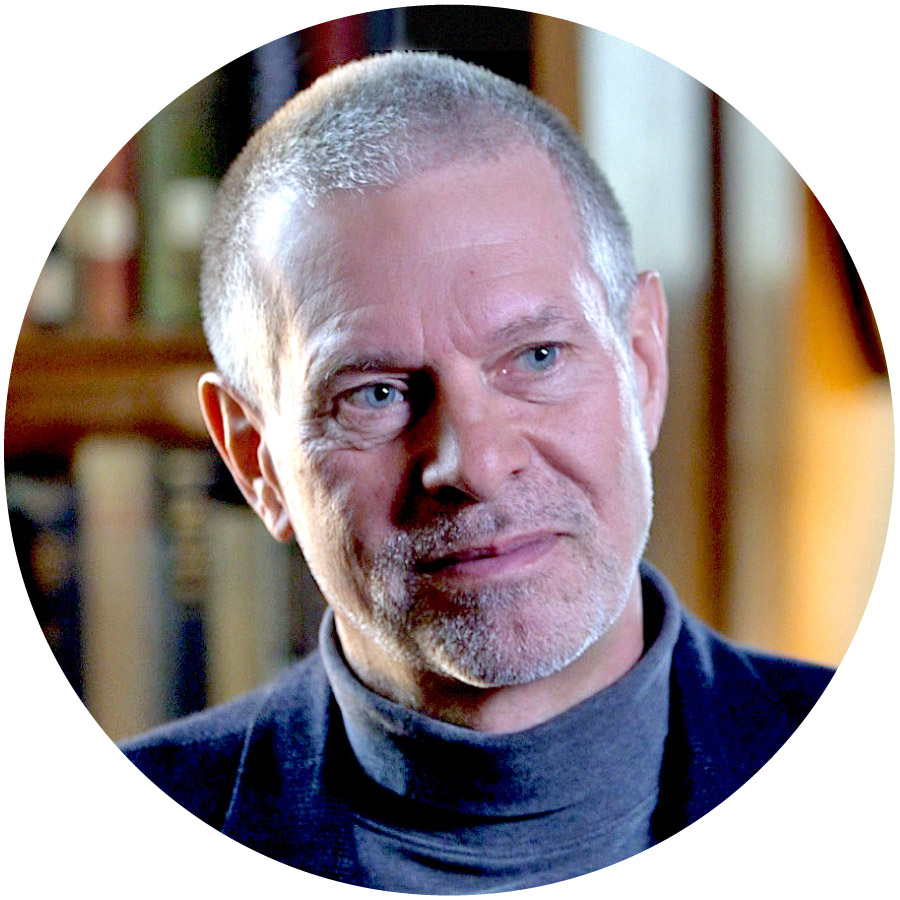 photo of a man with very short grey hair, a blue turtleneck, and a black blazer