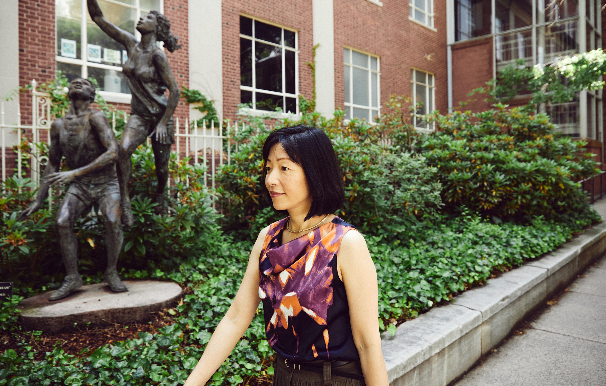 Akiko Iwasaki walks by a row of green bushes with bronze statues of children playing in the background