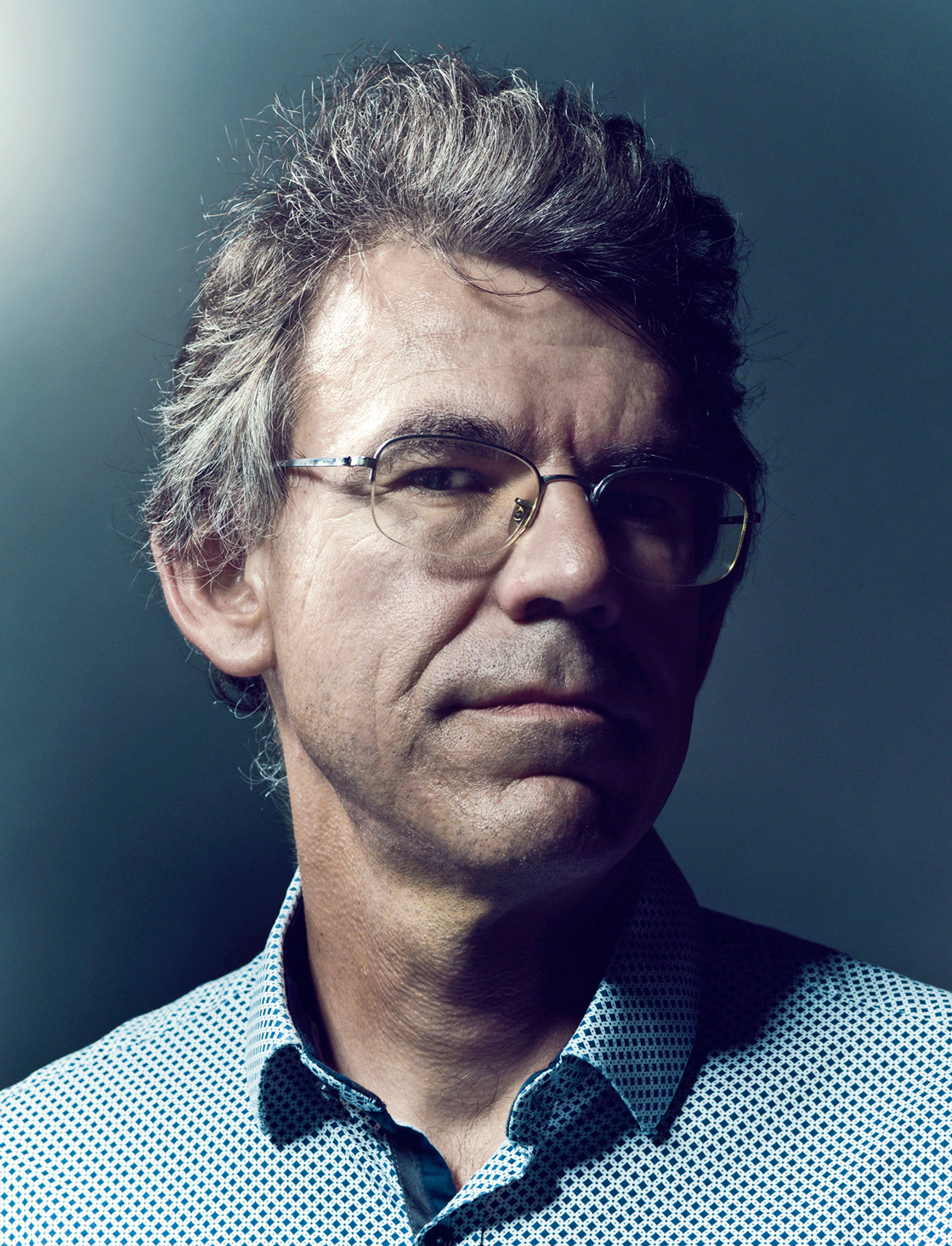 A close-up portrait of a bespectacled, serious, gray-haired man’s face.