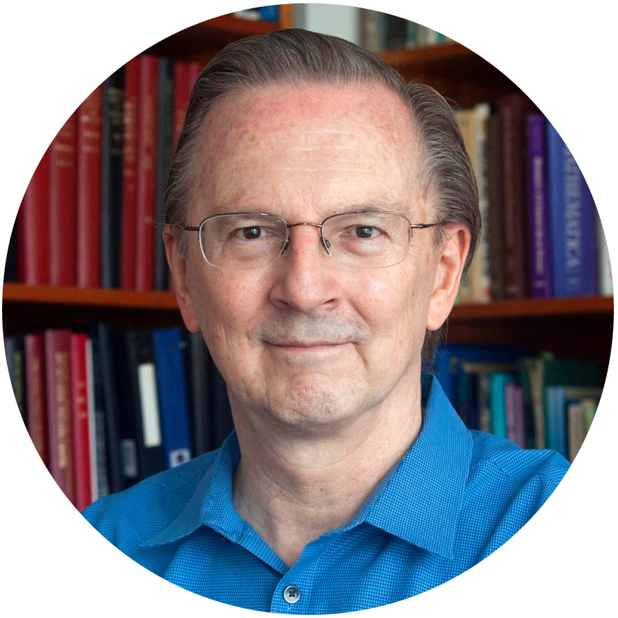 A man with light brown hair and glasses and a blue shirt smiles in front of a bookcase