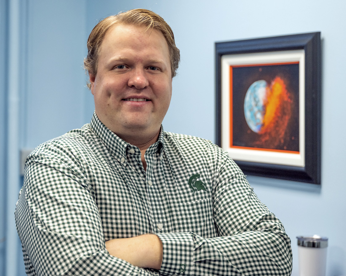 A blond man in a gingham shirt smiles at the camera. Behind him is a framed illustration of a planet undergoing an epic collision.