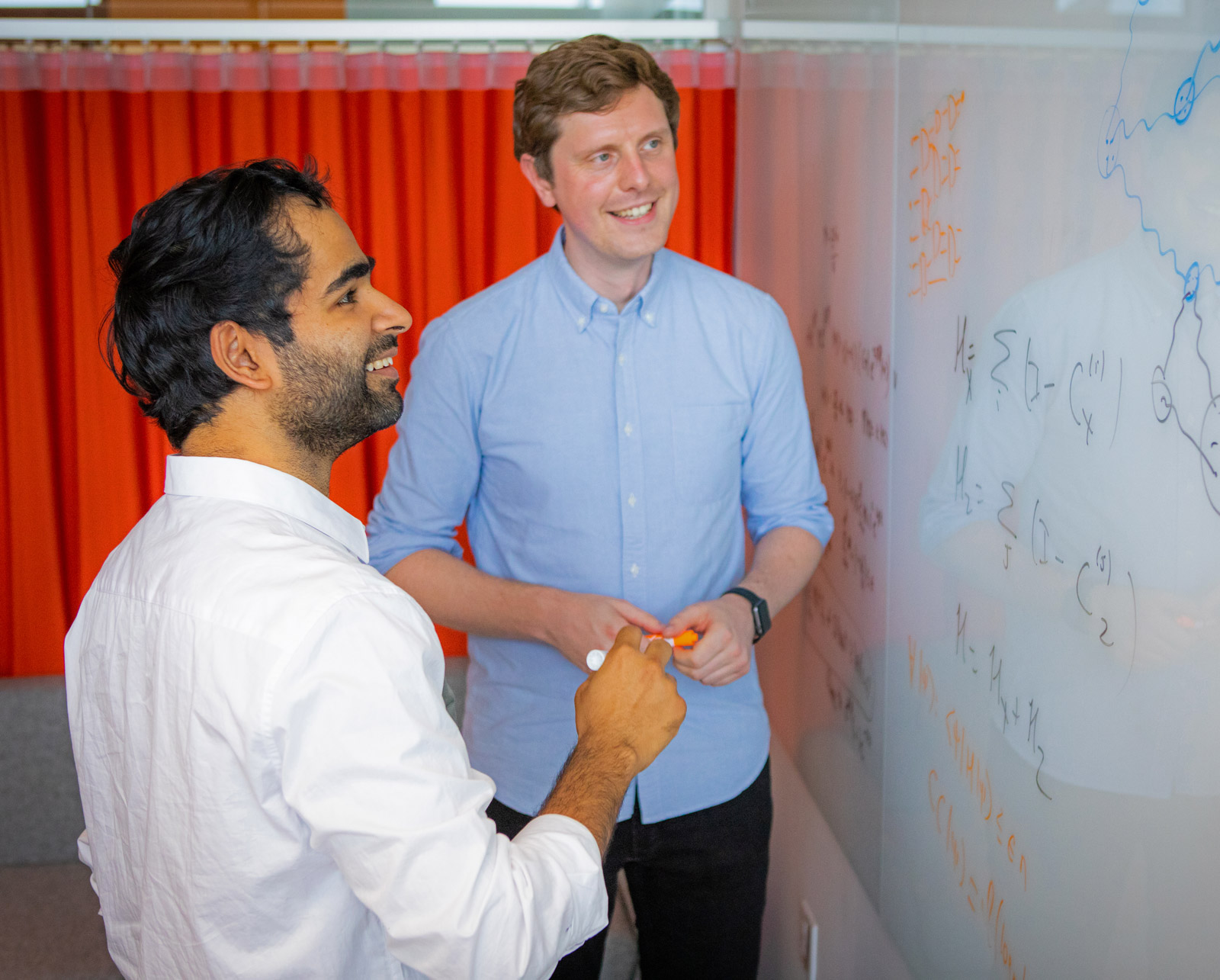 Anurag Anshu in a white shirt and Nikolas Breuckmann in a blue shirt look at equations on a white board.