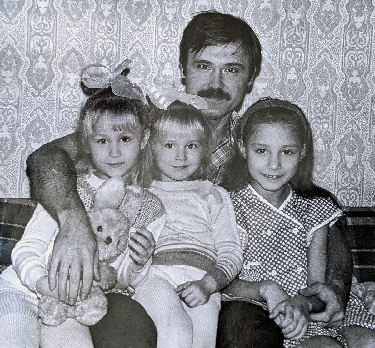 a black and white photo of a man with a mustache and three young daughters in front of patterned wallpaper