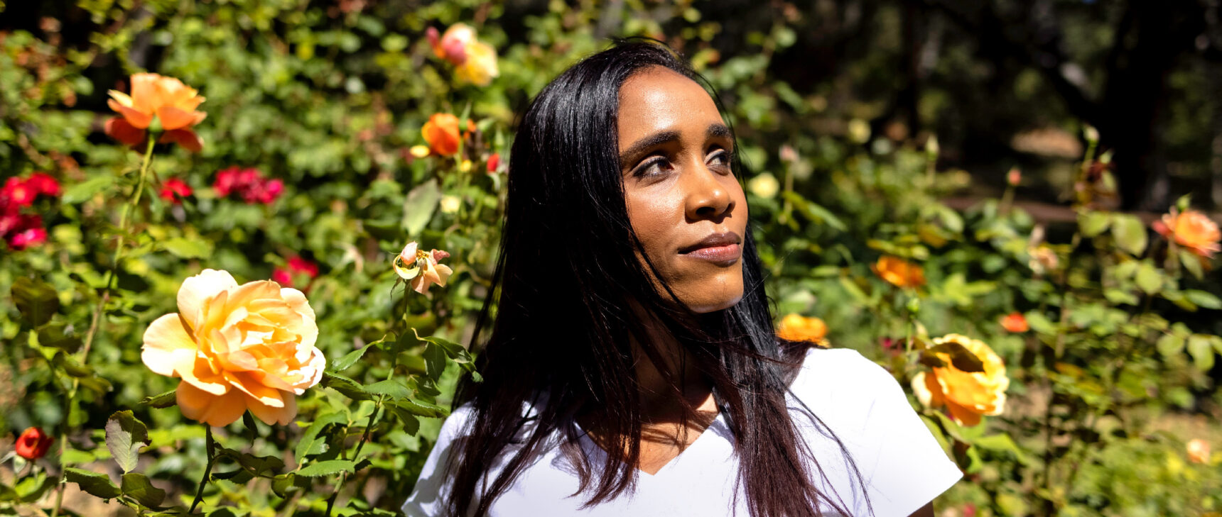 A woman in a white t-shirt is standing in front of a bush of orange roses, gazing to the side.