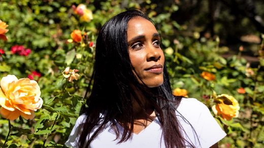 A woman in a white t-shirt is standing in front of a bush of orange roses, gazing to the side.