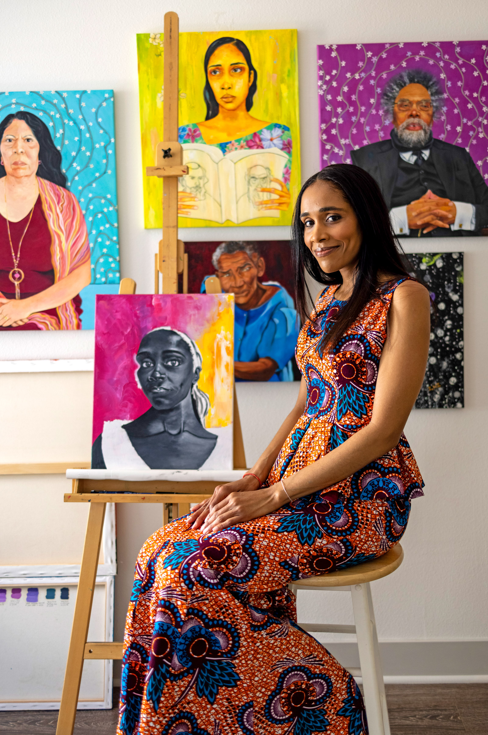 A woman in a West African print dress sits in front of several painted portraits hanging on a white wall