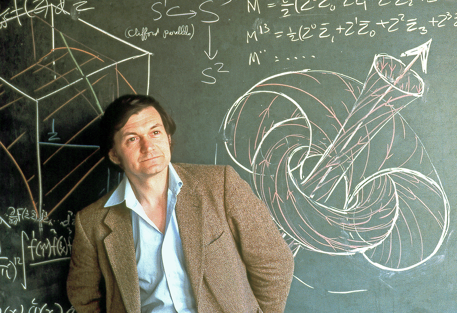 A man in a white shirt and brown blazer stands in front of a chalked-up blackboard, gazing into the distance.