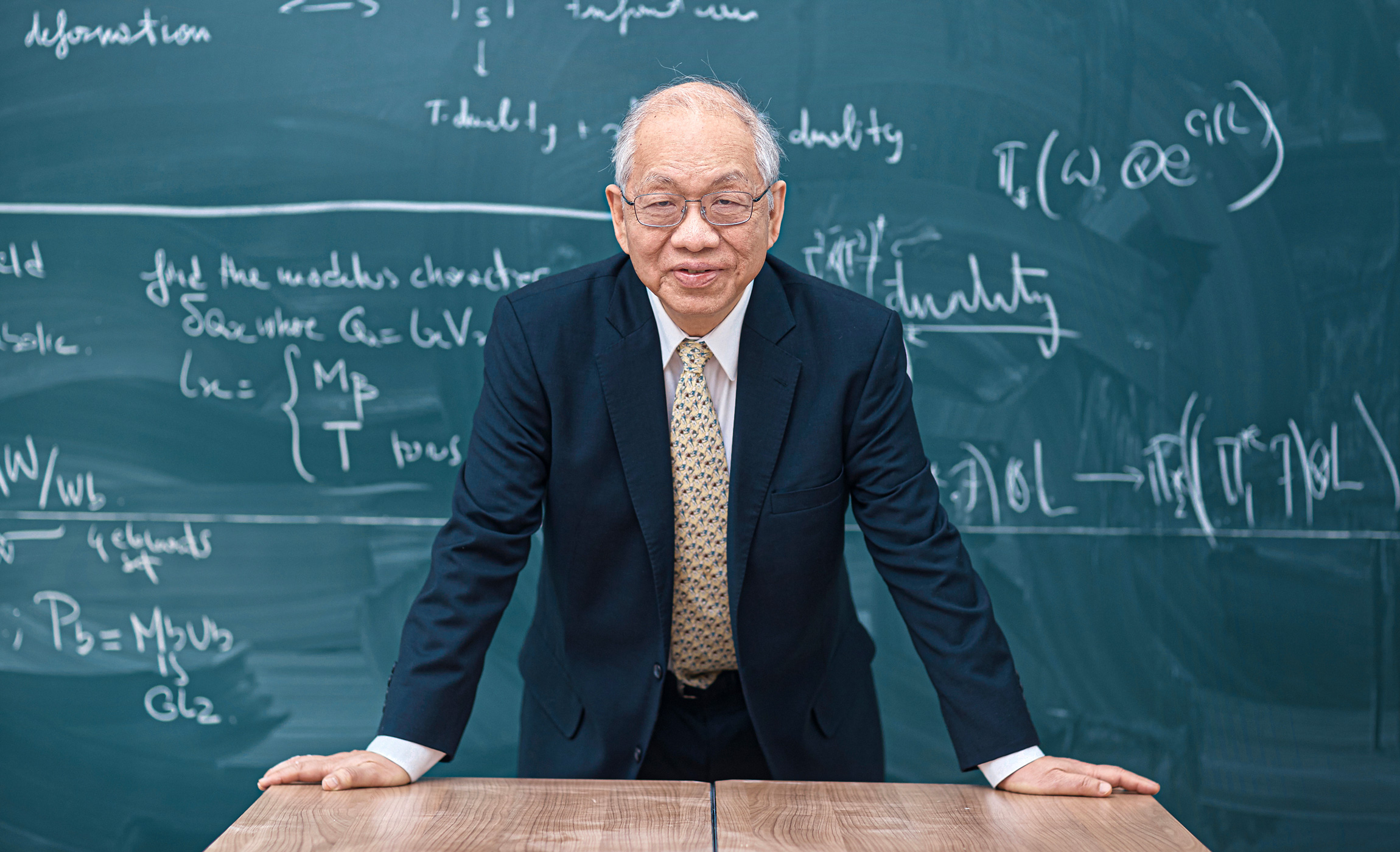 A portrait of Shing-Tung Yau in a gray suit and yellow tie standing in front of a chalkboard