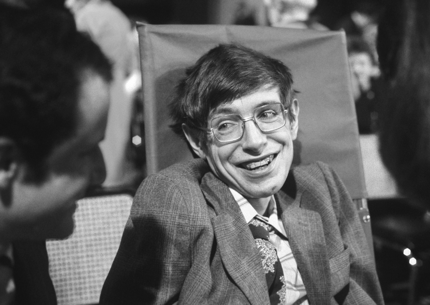 A black-and-white photo of a young Stephen Hawking in a wool suit, seated and smiling at a colleague