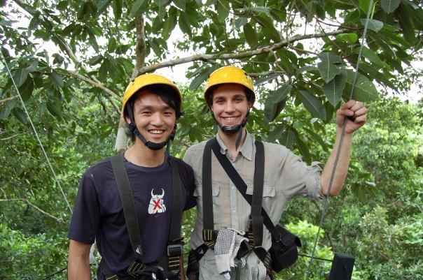 Two men wearing hard hats and safety gear smile at the camera from a platform in the canopy of the Amazon rainforest.