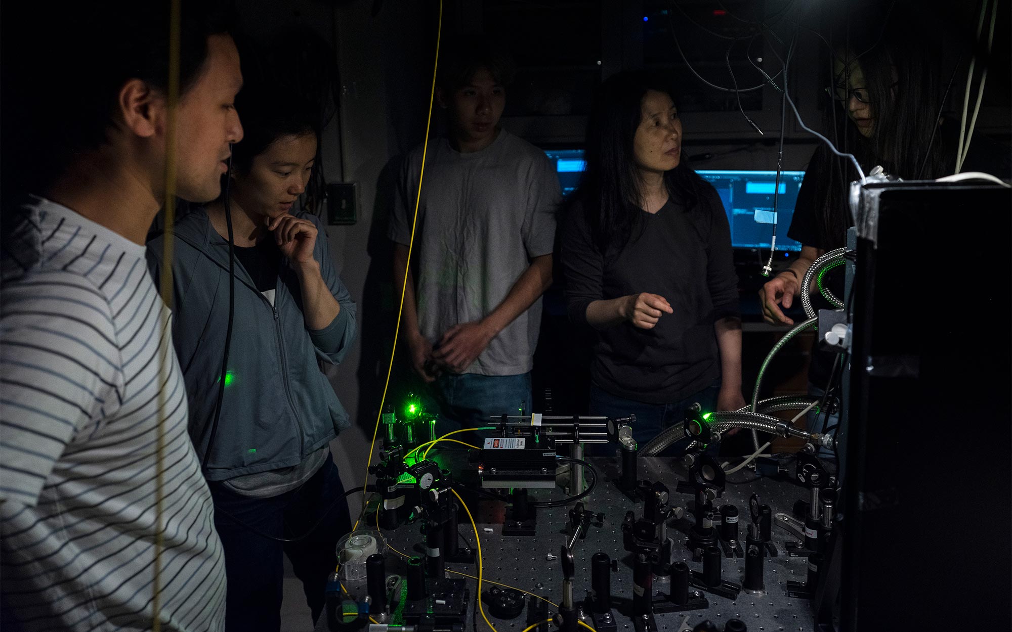 A photo of several researchers, including Shan and Mak, standing in a dark room around an optical table illuminated by laser light.