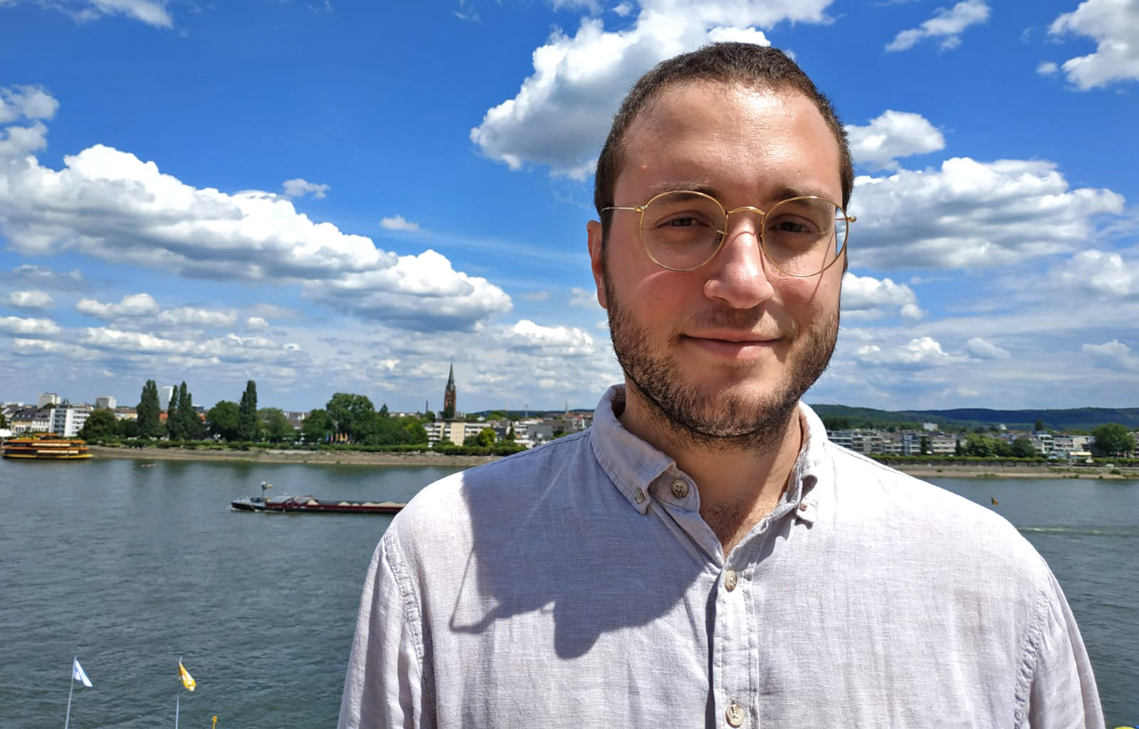 photograph of a light-skinned man standing in front of a river with a small city and trees in the background. The sky is bright blue and full of clouds, and the man has brown hair, a short beard, and is wearing gold-rimmed glasses and a blue collared shirt.