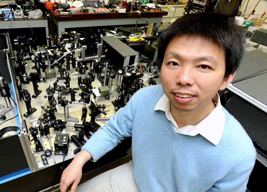 A man in a blue sweater stands in front of an optical table.