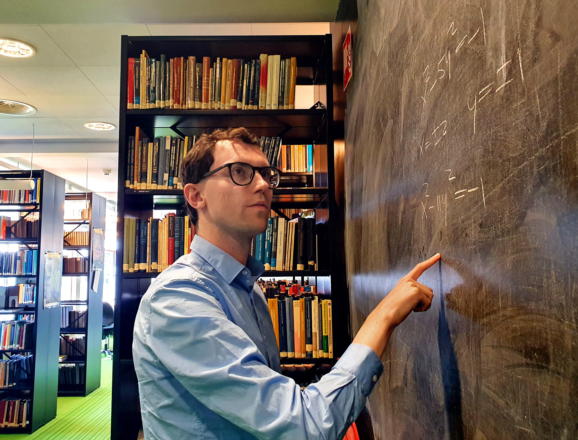 A photograph of a light-skinned man pointing at an equation on a blackboard in a library. He has short brown hair, black-rimmed glasses, and is wearing a blue collared shirt.