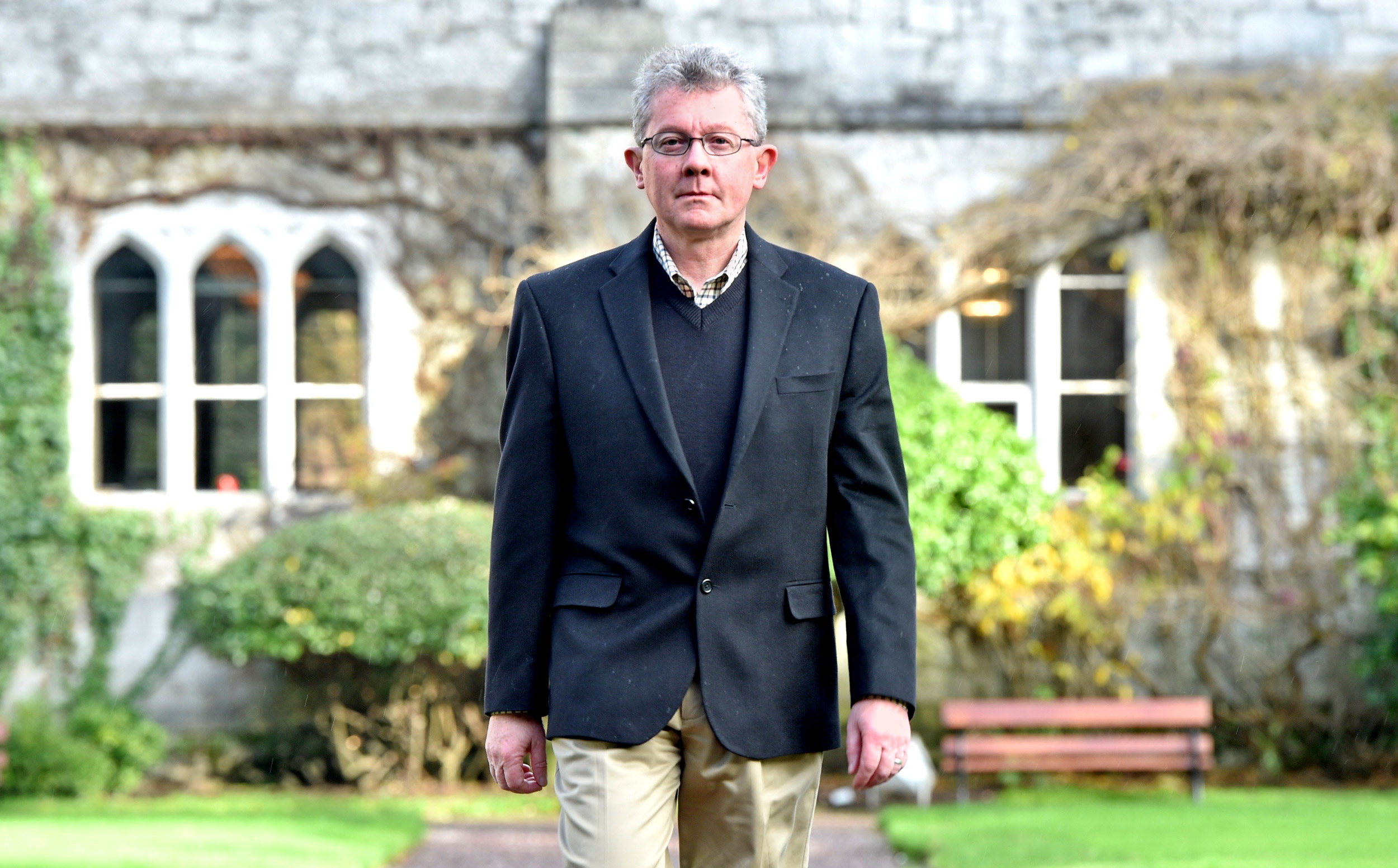 A man in a blazer with curly gray hair and glasses walks toward the camera in front of an ivy-covered stone building.]